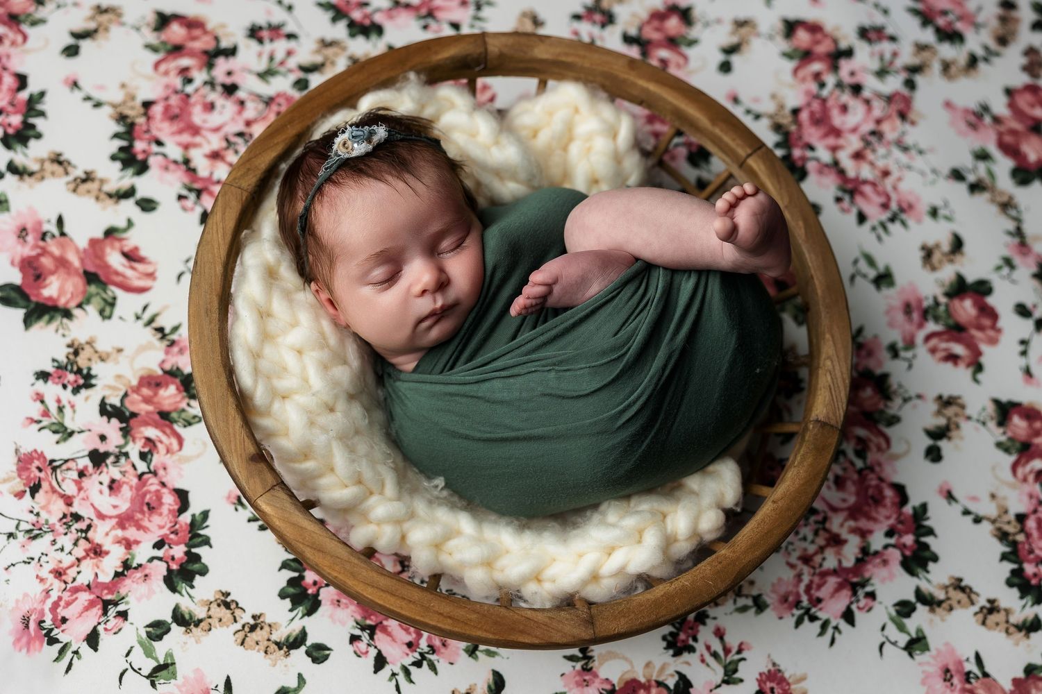 Sleeping infant wrapped in green swaddle resting in round wooden bowl on cream cushion with floral background.