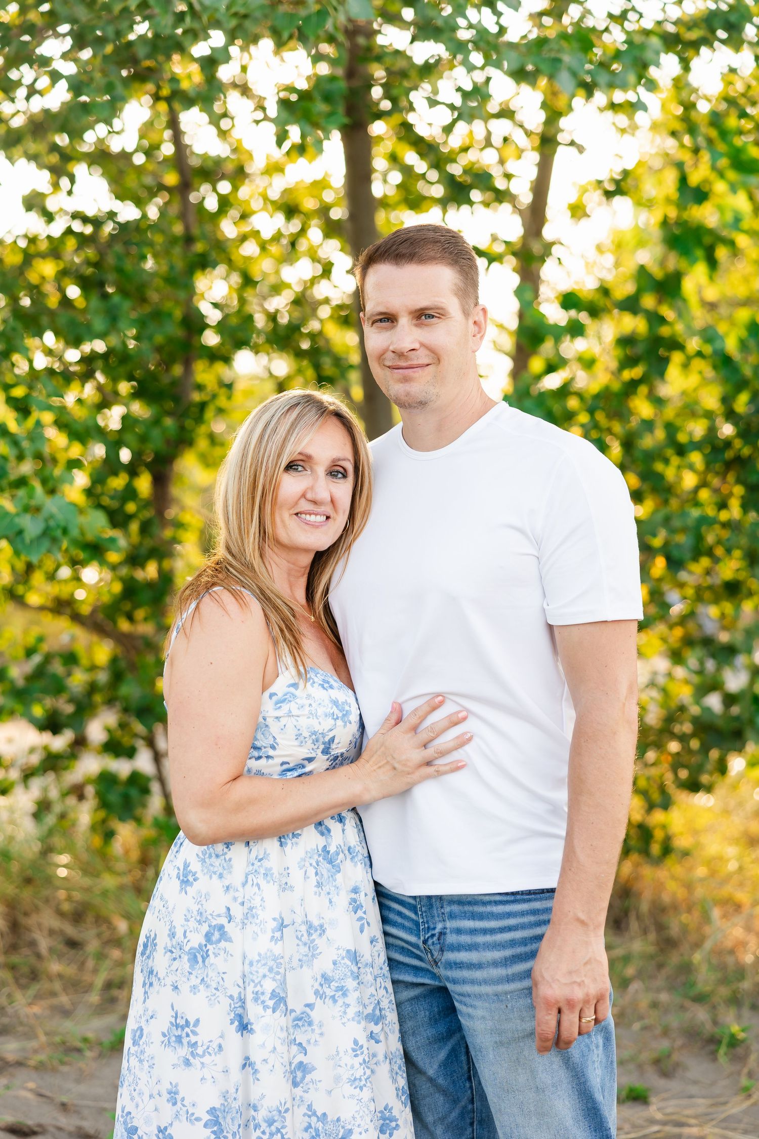 A couple poses outdoors on a sunny day, with green trees and golden sunlight in the background.