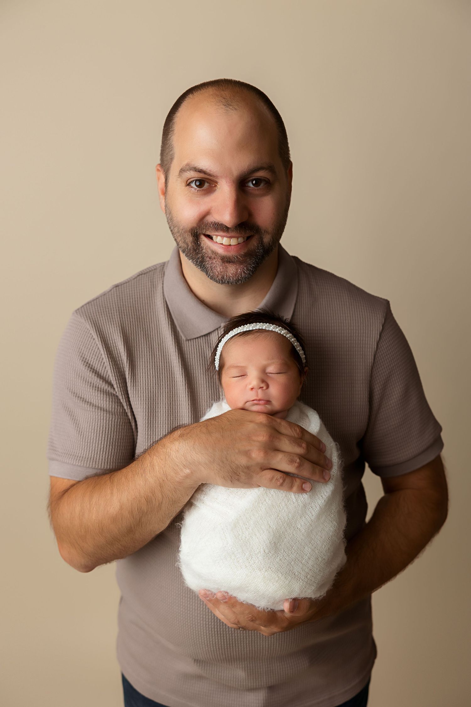 A dad in a grey shirt holds his sleeping newborn wrapped in a white blanket against neutral background.