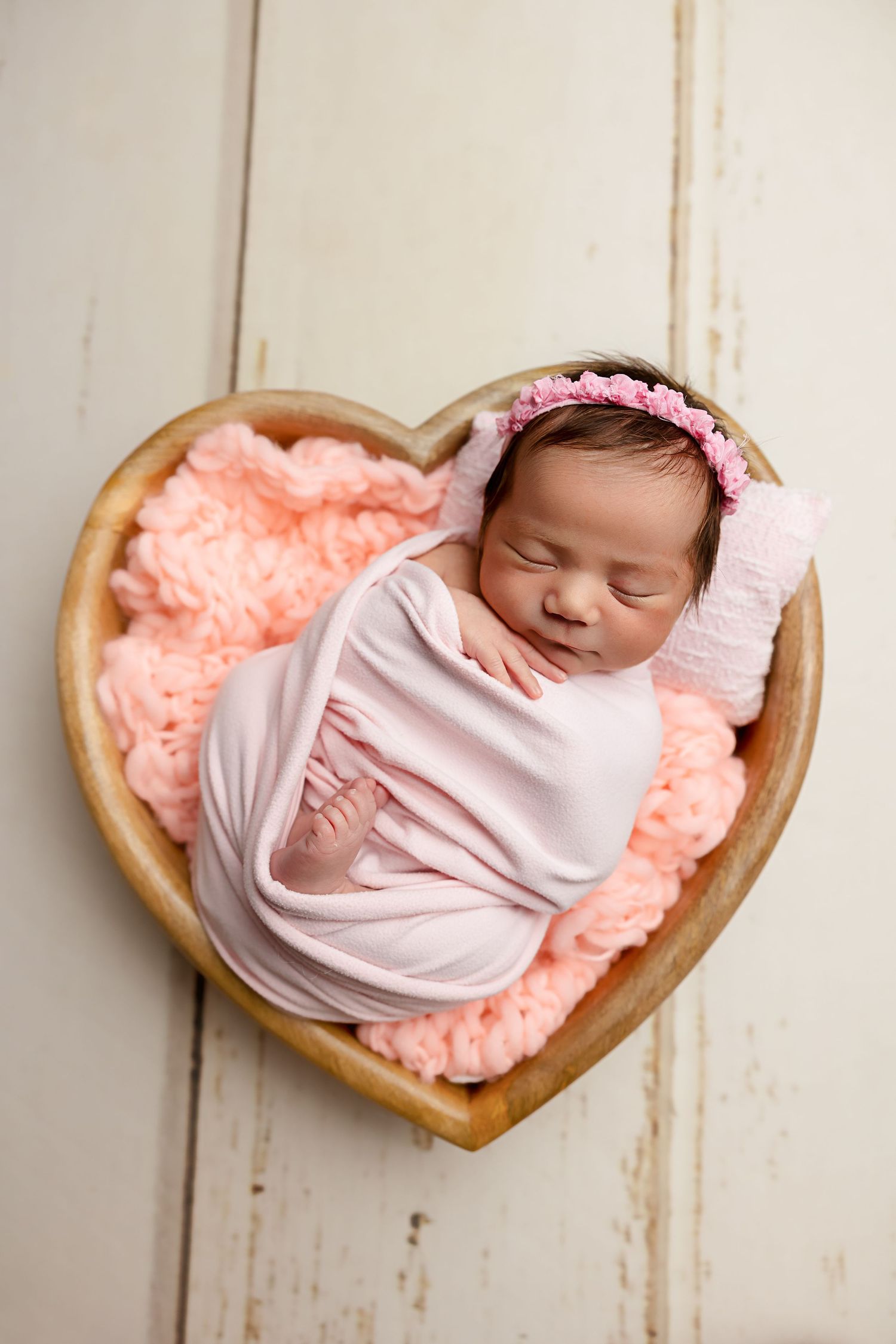 Newborn sleeping peacefully in heart-shaped wooden bowl lined with pink fur padding.