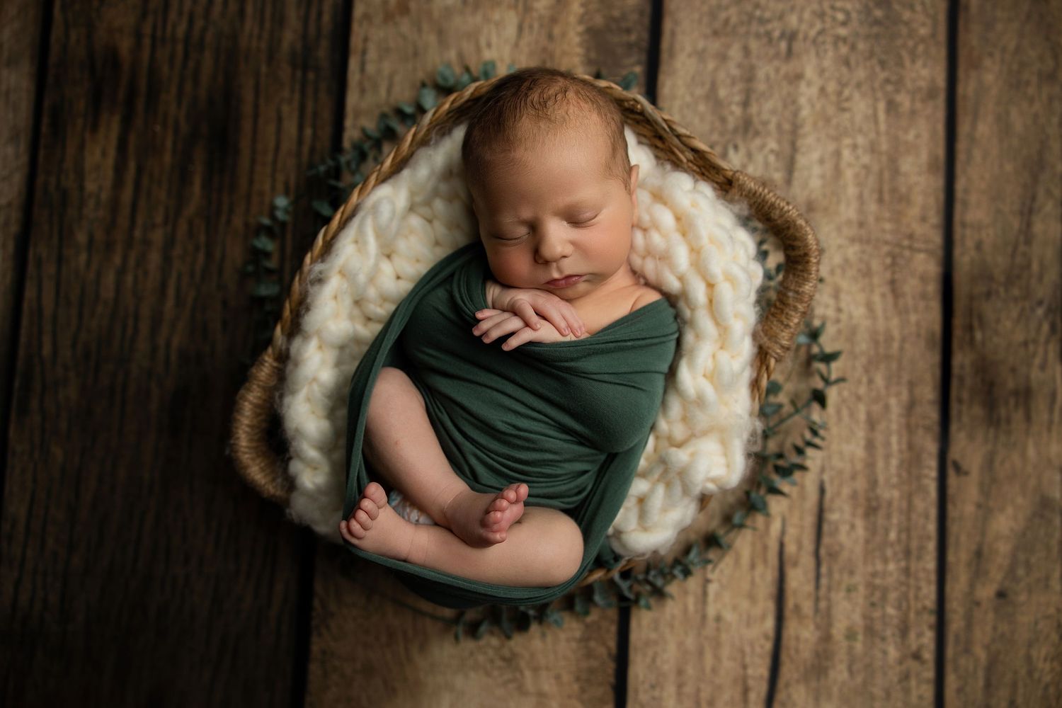 Newborn resting in green wrap inside cozy round basket on wooden floor.