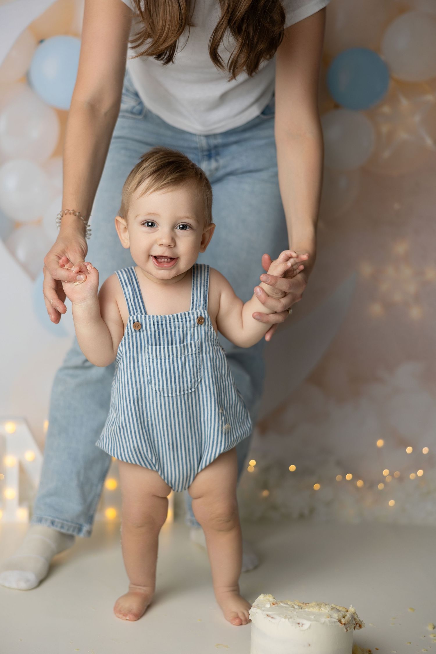 First birthday photoshoot with supportive hands, blue striped outfit, and festive balloon backdrop.