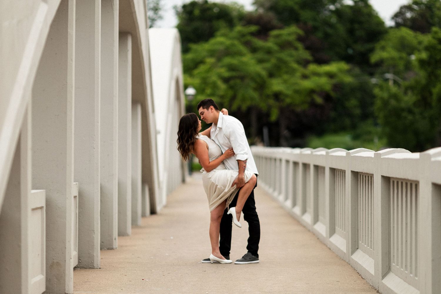 Couple sharing a romantic kiss on a white bridge walkway surrounded by lush green trees.
