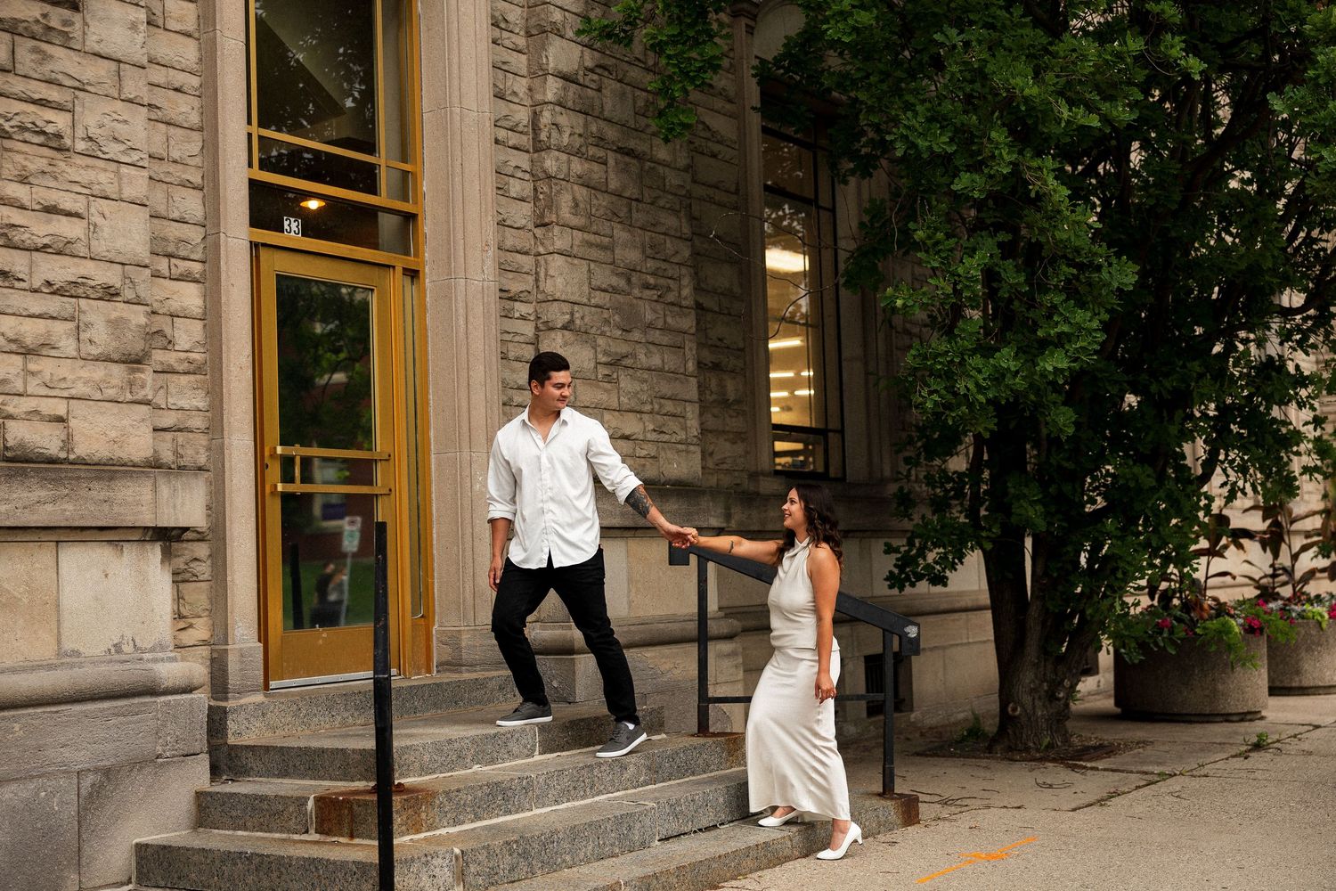 Couple holding hands on urban building steps with green trees nearby.