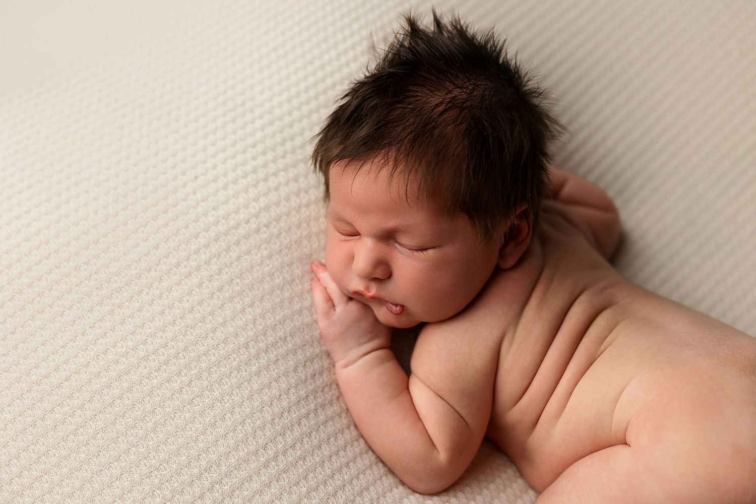 Sleeping newborn baby curled up on soft white textured blanket in natural light.