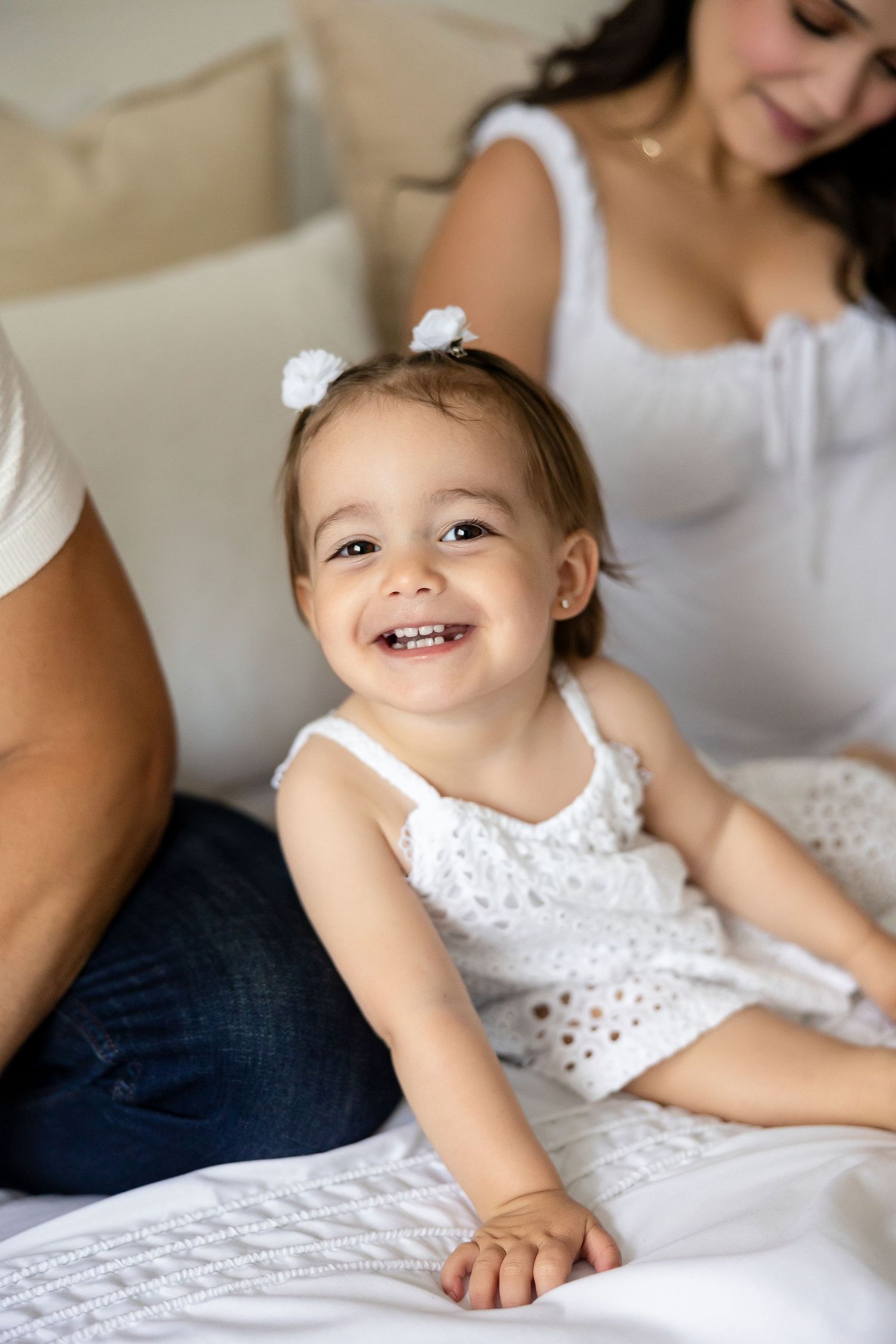 Sweet candid portraits show a child in a white lace dress lying on white bedding with joyful expressions.
