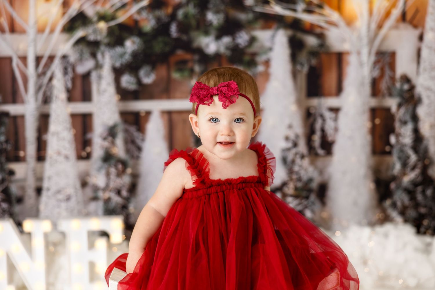 Small child in flowing red dress stands against winter forest backdrop with white trees and lights.