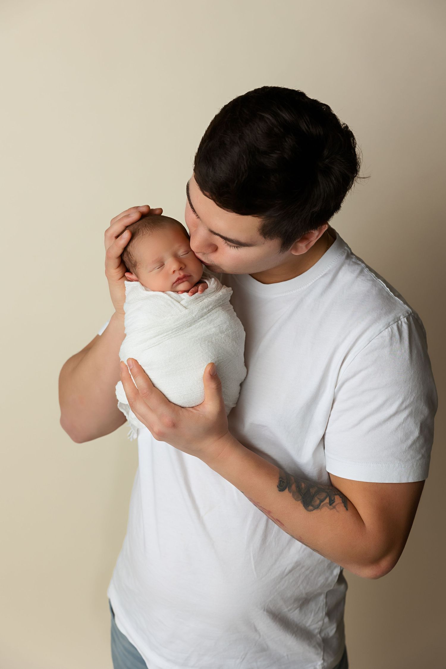 Parent tenderly kisses sleeping newborn baby wrapped in white swaddle.