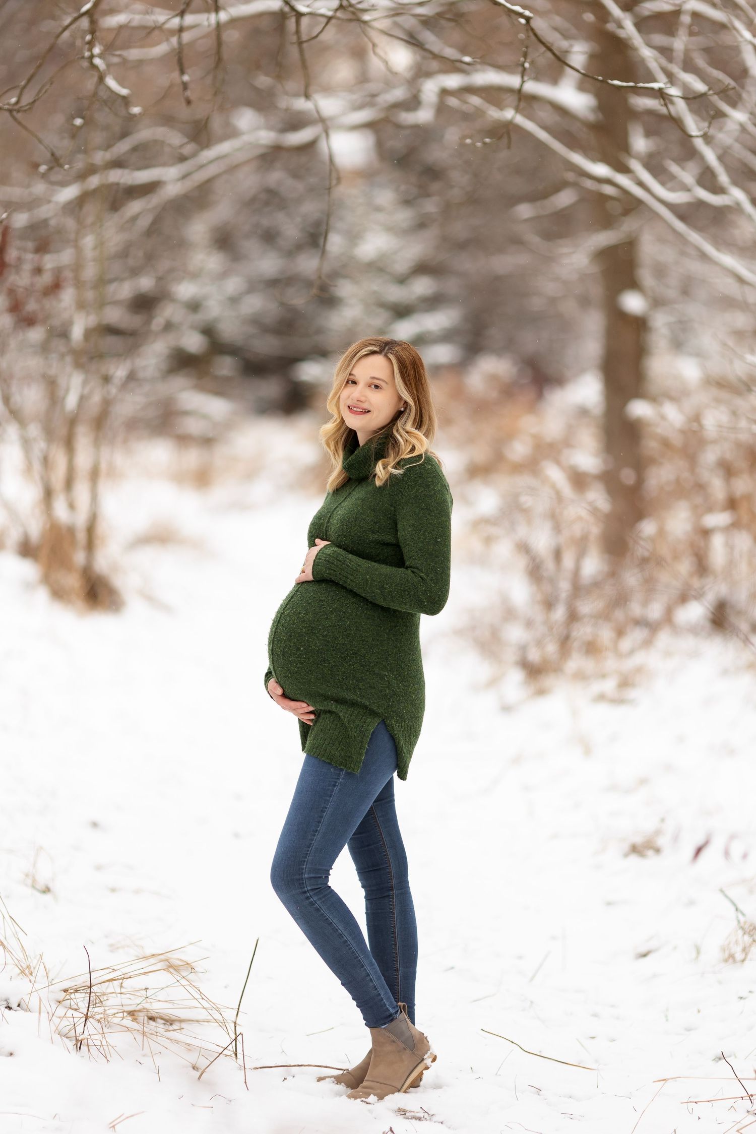 A pregnant figure stands in a snowy winter scene wearing a green sweater and jeans.