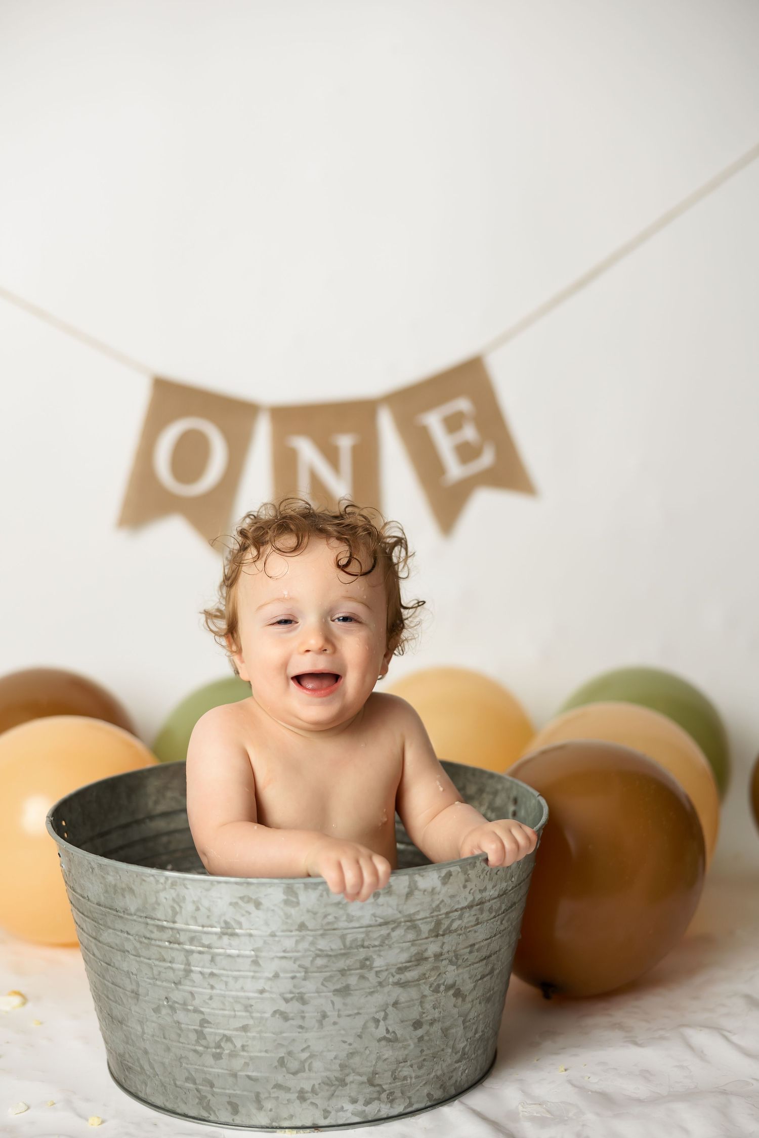 Happy baby sitting in metal tub with ONE banner and colorful balloons in background.