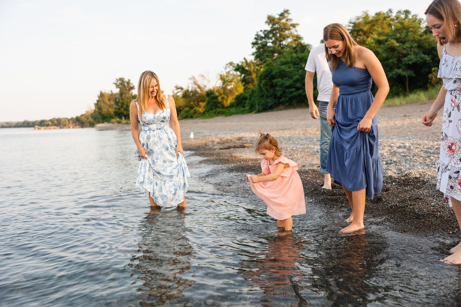 Several people wearing summer dresses stand at the edge of a lake during sunset, dipping their feet in the water.