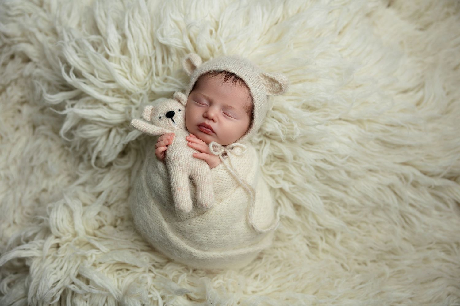 Sleeping newborn wrapped in cream swaddle laying on fluffy white fur backdrop.