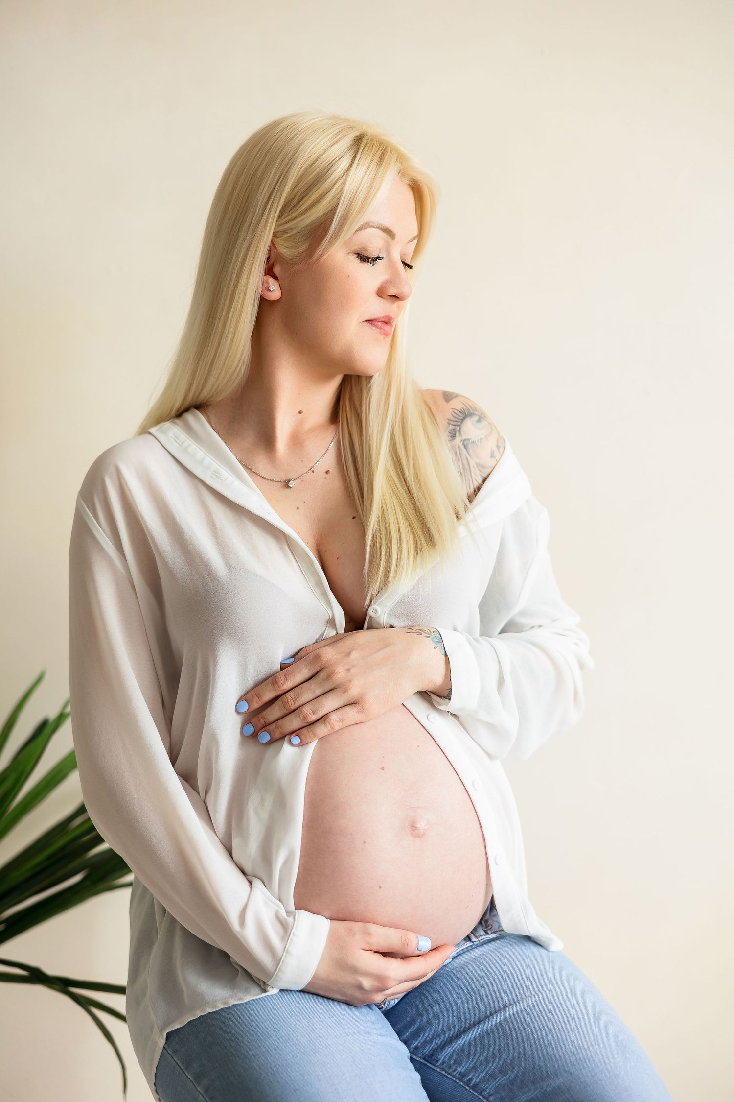 A serene maternity portrait against a cream wall with indoor plants providing natural decor.