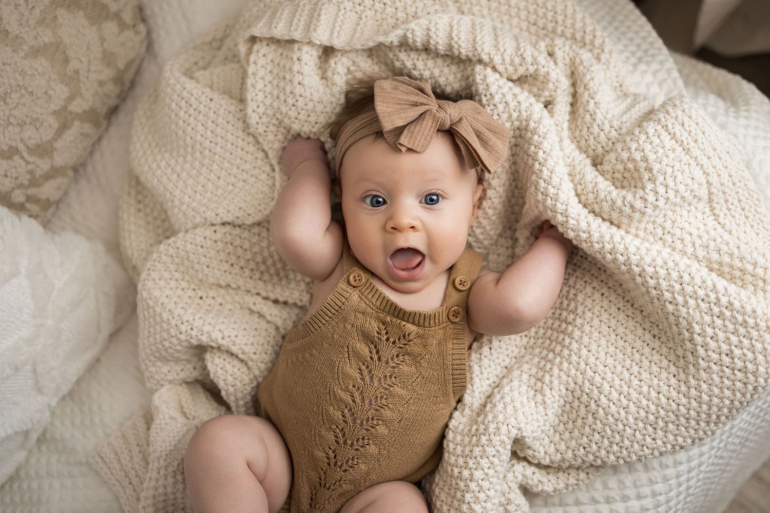 Adorable baby in knit brown romper with bow headband, lying on cream textured blanket with mouth open in surprise.