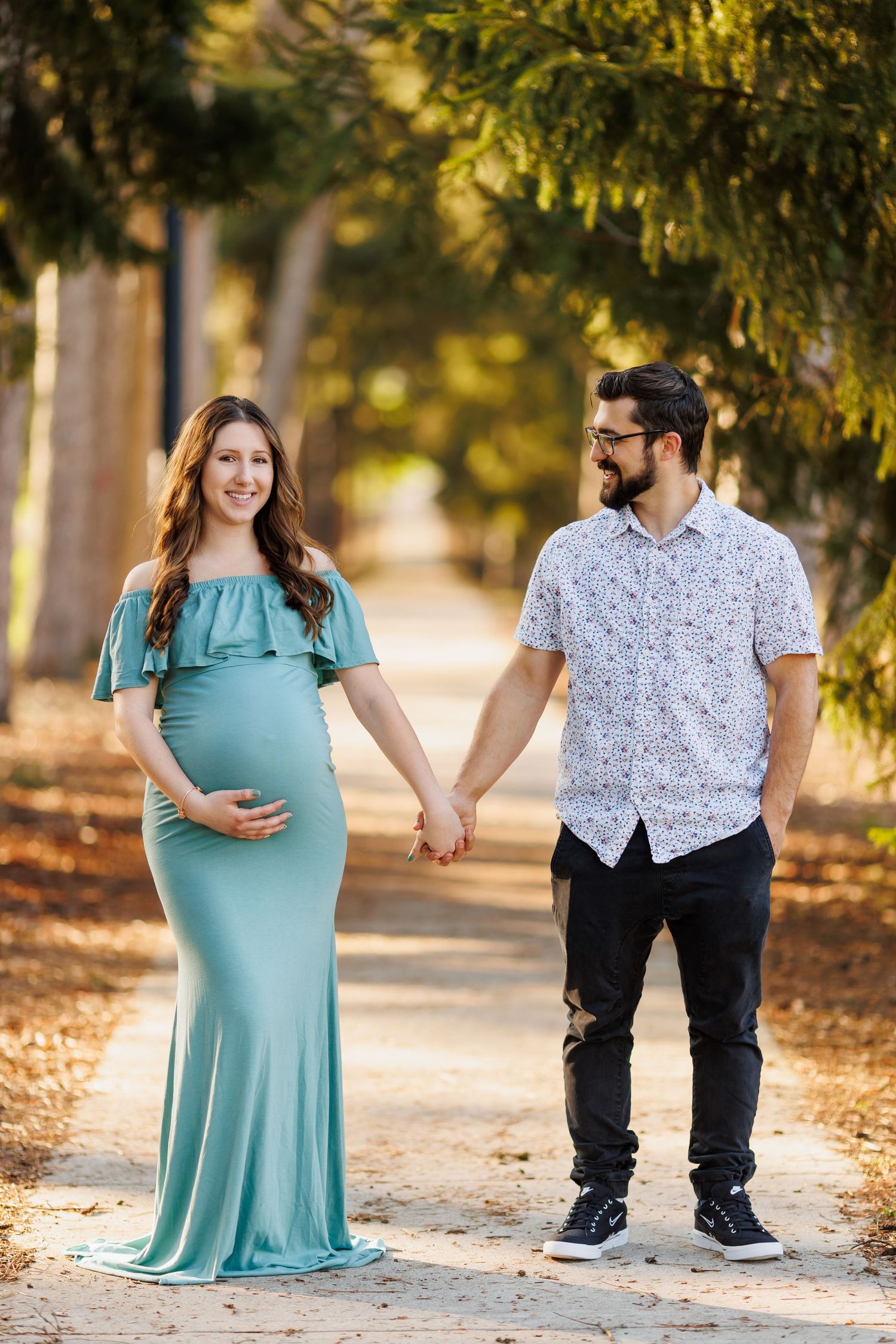 Outdoor maternity session with couple in teal dress and patterned shirt on tree-lined path.
