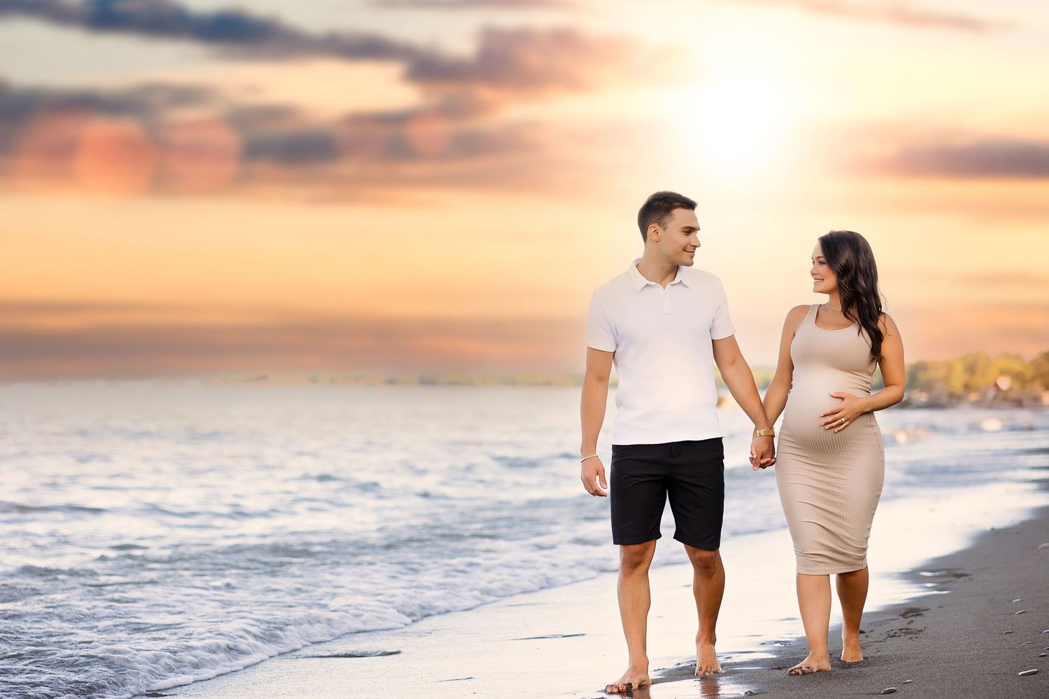 Couple walks hand in hand on beach during sunset maternity photoshoot.