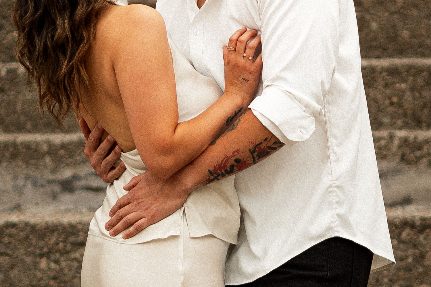 Couple embracing tenderly, woman in white dress, man with floral tattoo on arm, romantic outdoor setting.