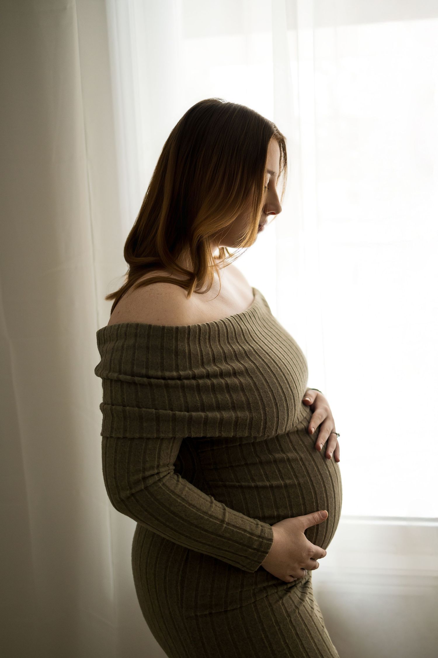 Artistic maternity photo series showing silhouette against window light in cozy sweater.