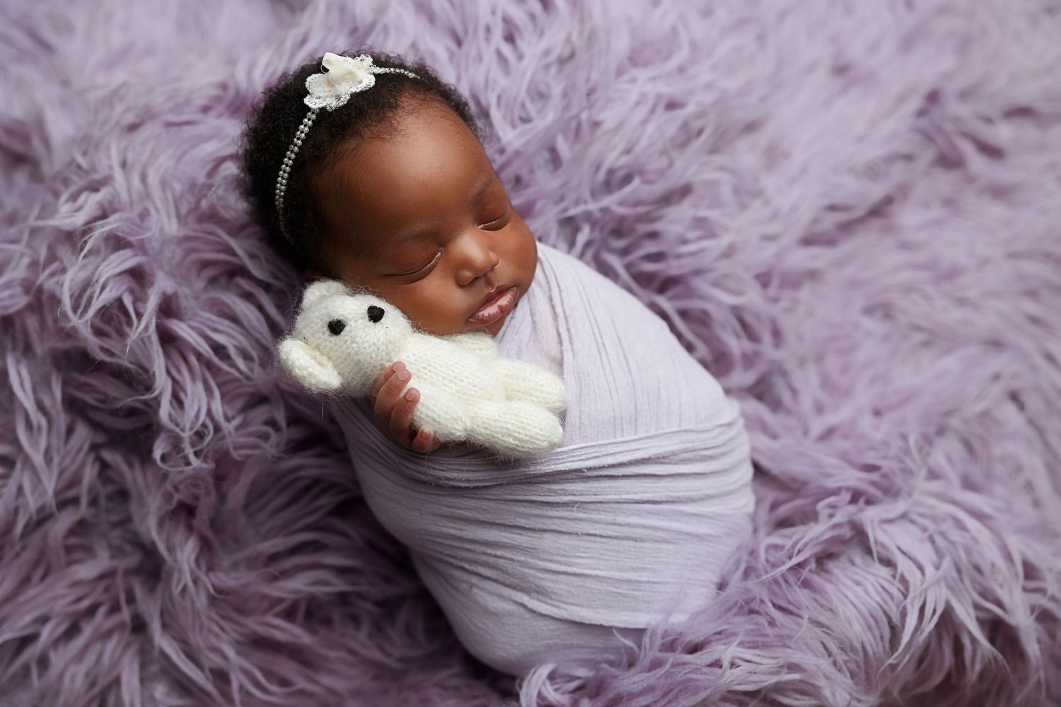 A newborn baby wrapped in gray fabric lies on a fluffy purple blanket with a small white plush toy.