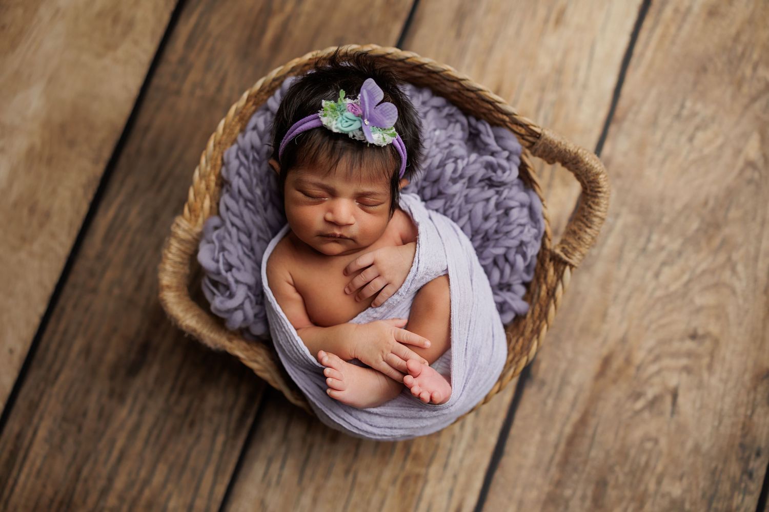 A sleeping newborn baby wrapped in purple fabric lies peacefully on a rustic wooden floor.