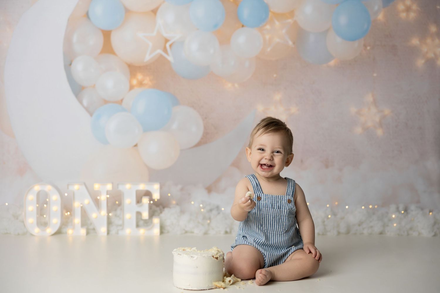 First birthday setup with blue and peach balloon garland, twinkle lights, and illuminated ONE sign.