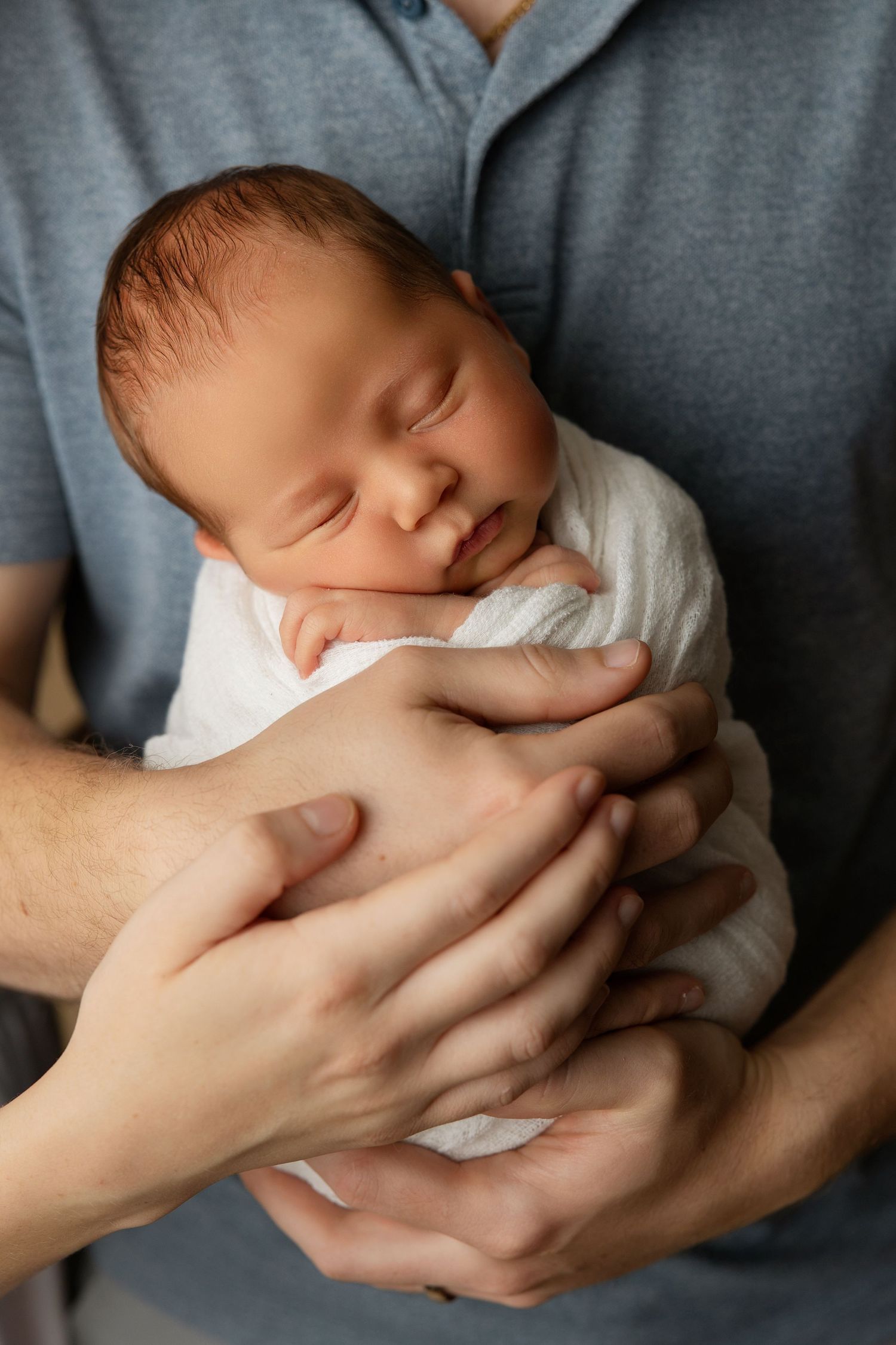 A sleeping newborn baby swaddled in white being cradled gently in adult hands against a gray shirt.