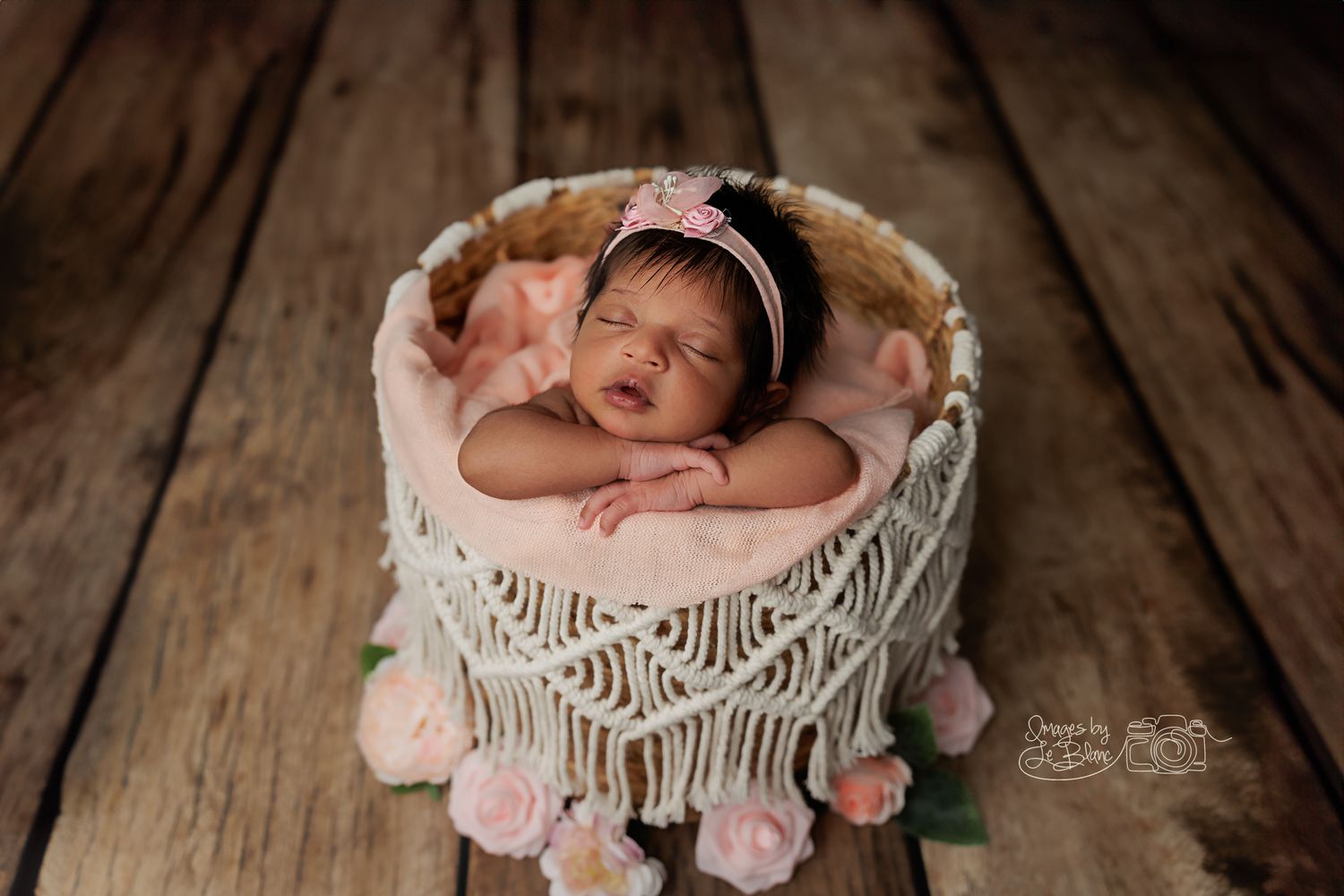 Newborn resting in macrame basket with pink pom pom decorations on wooden floor.