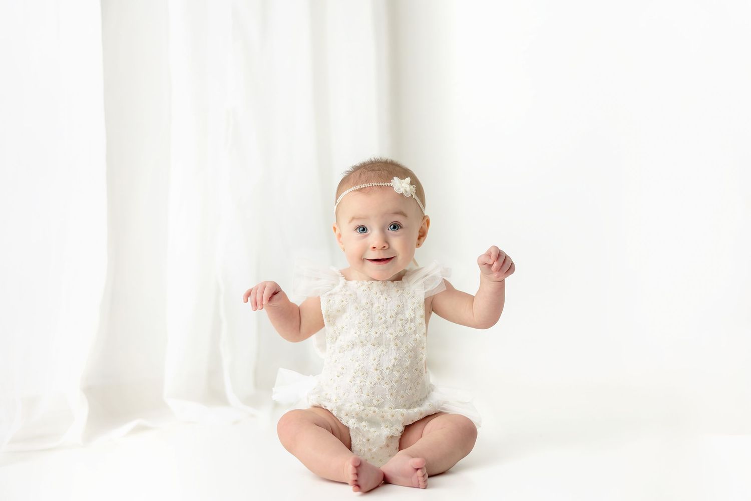 Baby in white lace outfit photographed against bright white backdrop.