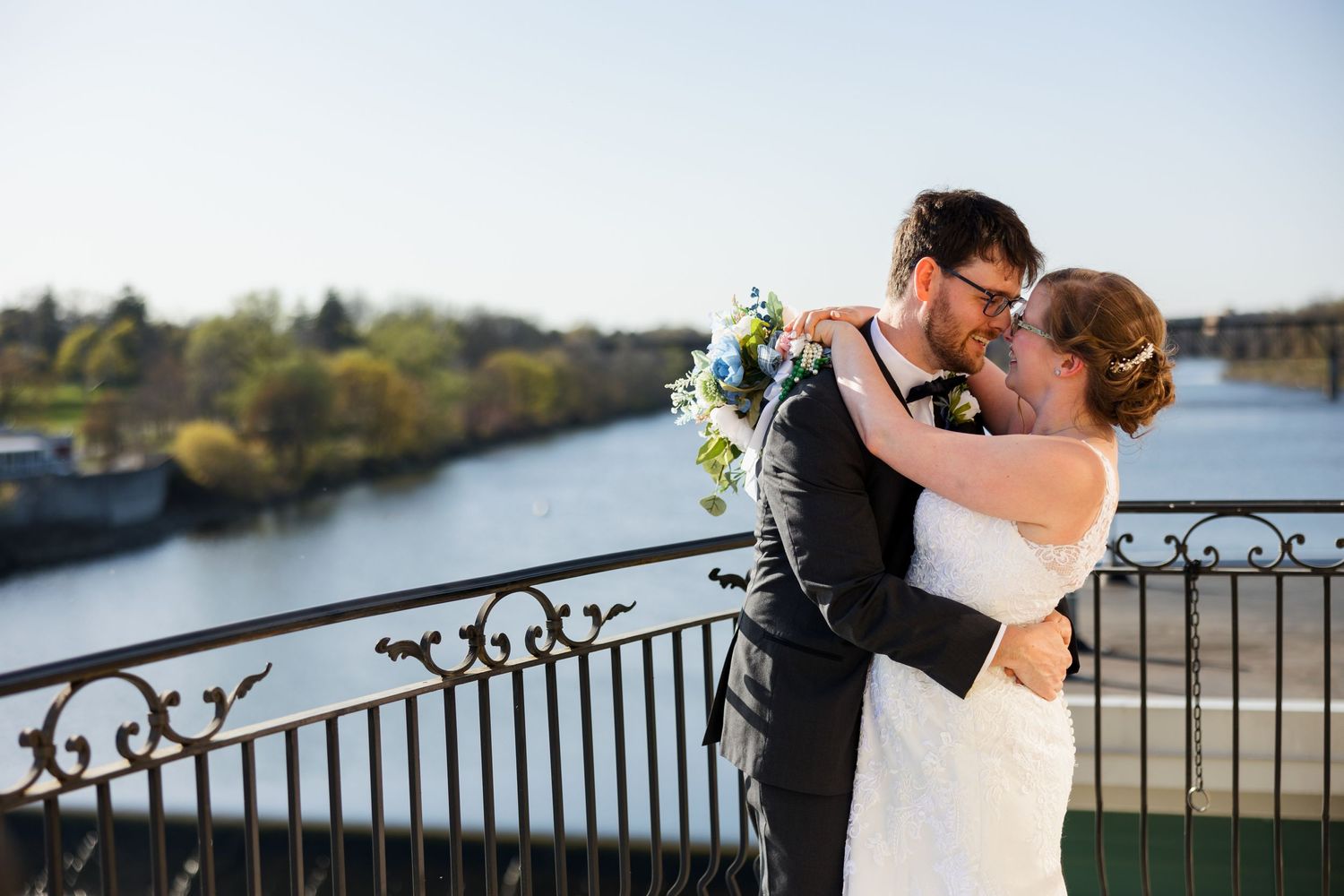 Couple embracing on balcony overlooking river during sunset wedding photoshoot.