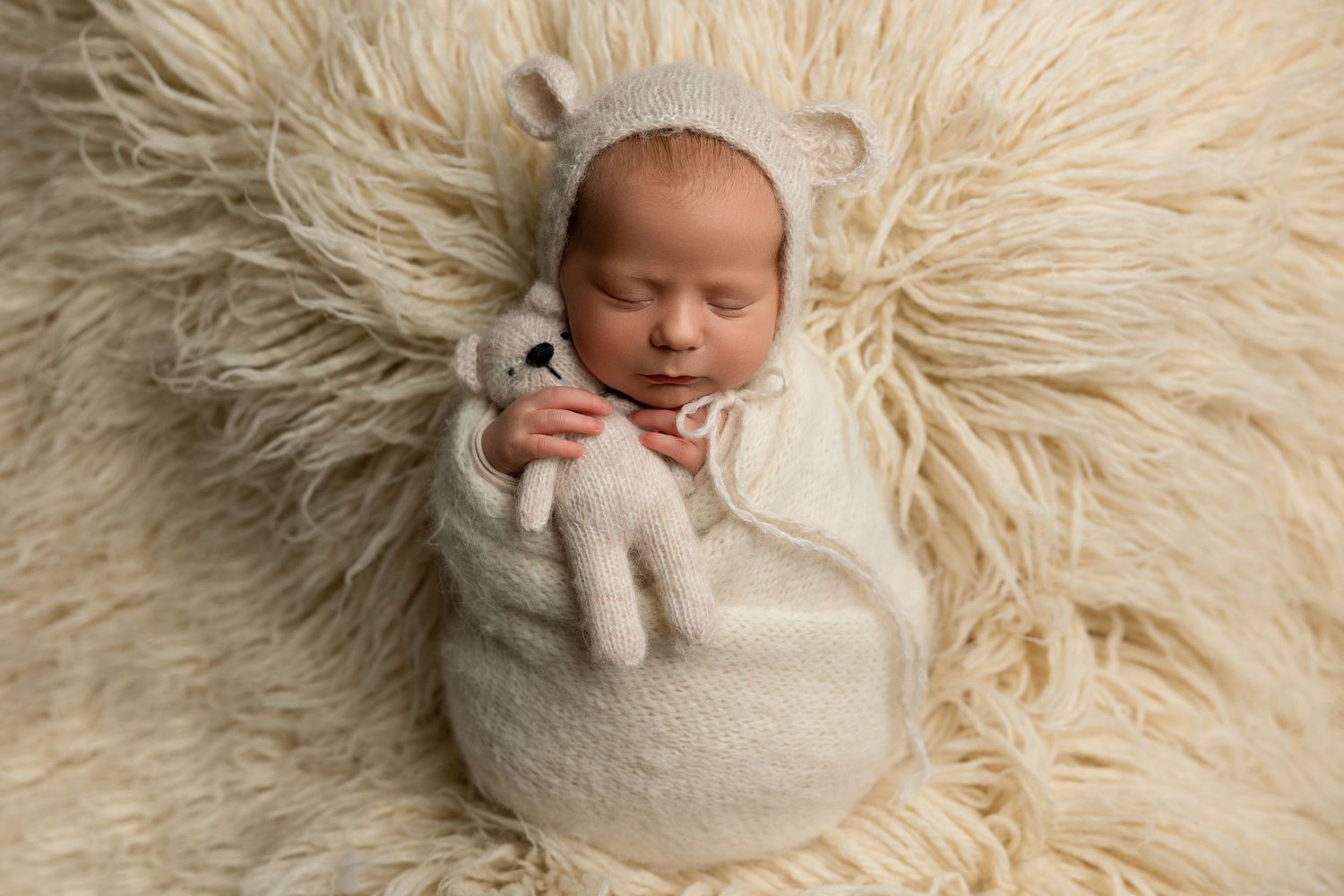 Sleeping newborn wearing bear ears bonnet on fluffy cream textured backdrop with stuffed animal.