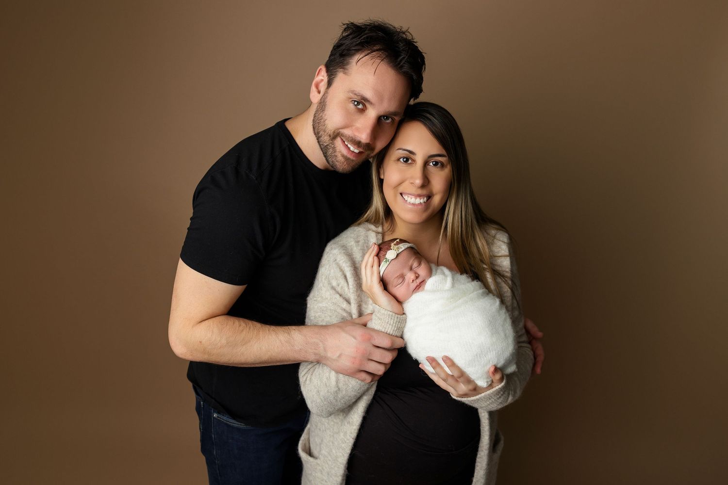 Studio family portrait with newborn baby against brown backdrop.