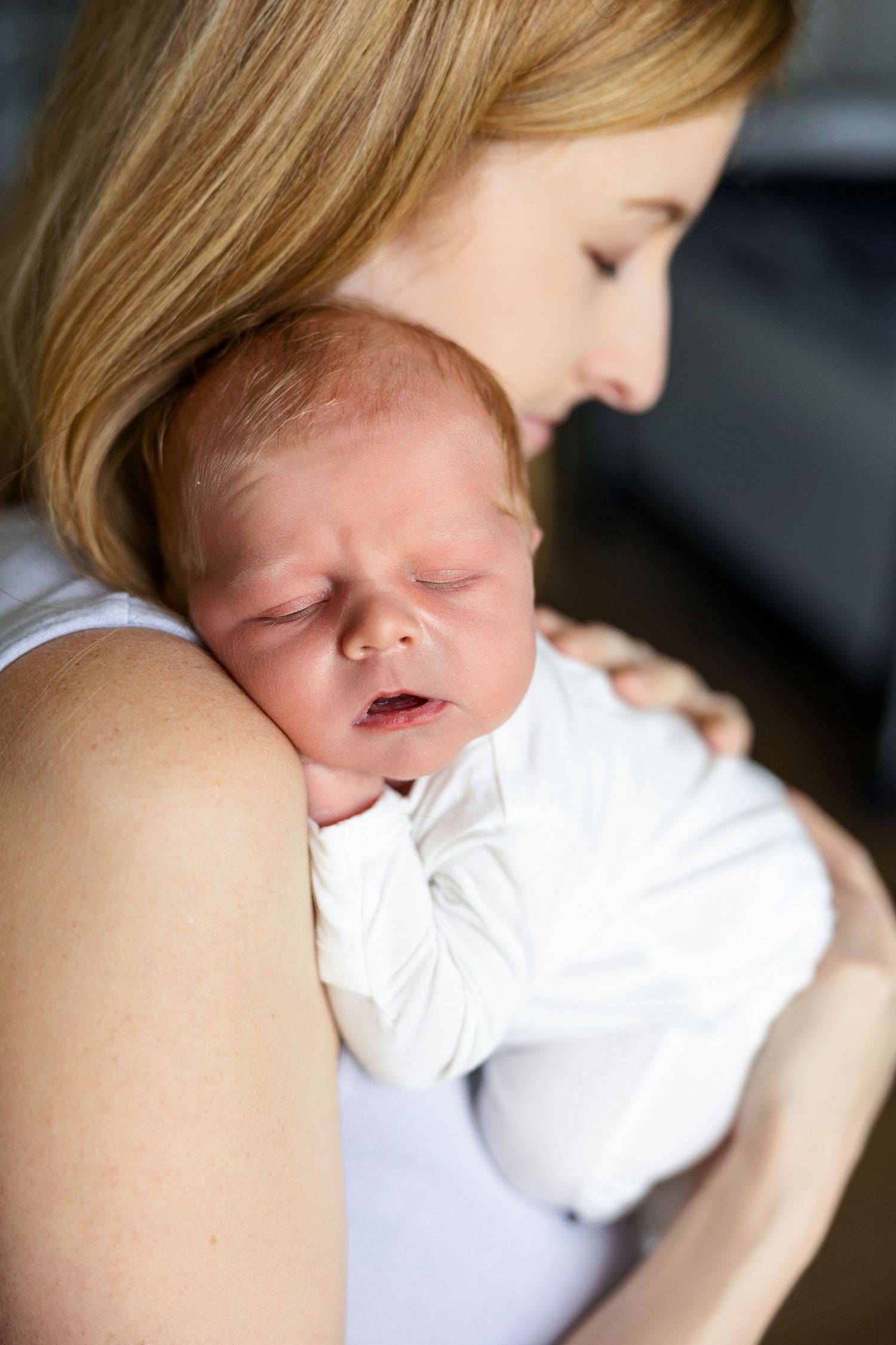 A tender moment shows a sleeping newborn baby in white clothing cradled against someone in soft natural lighting.