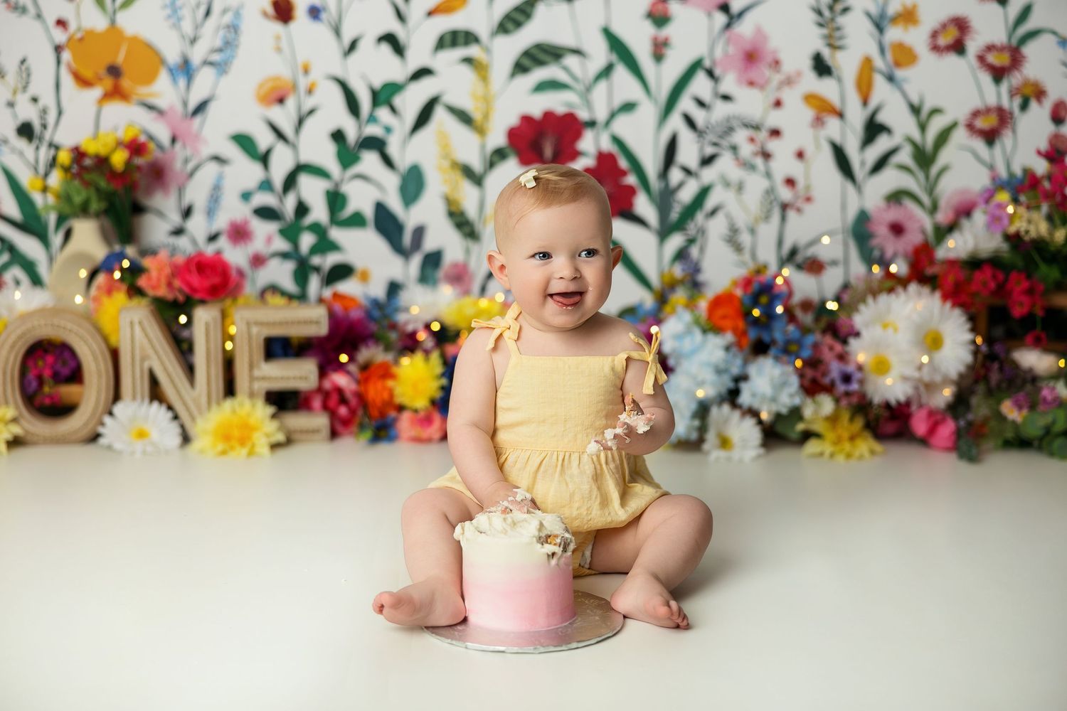 Baby girl in yellow romper sits smiling before a floral backdrop with ONE letters and pink smash cake for her first birthday.