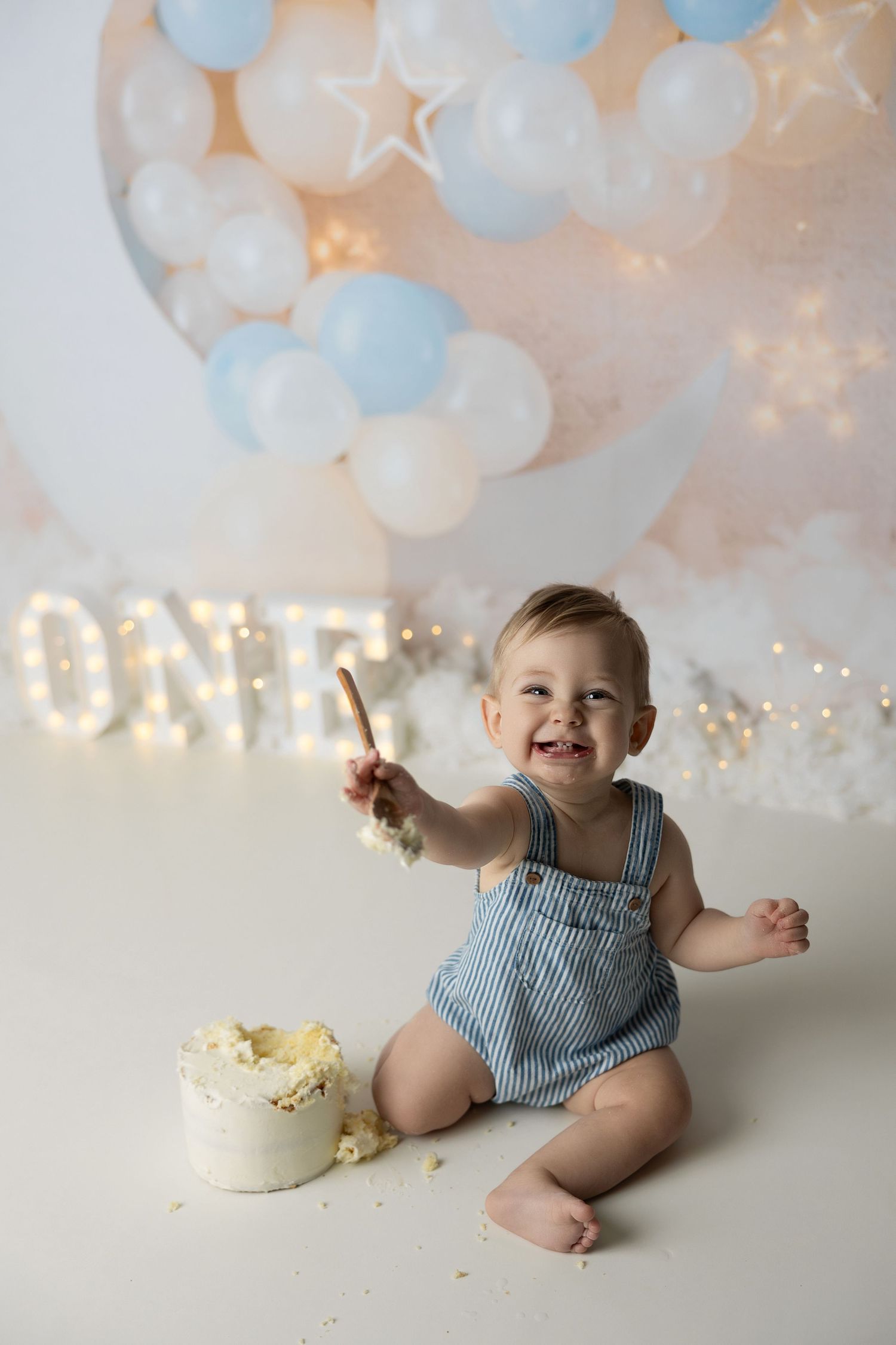 Sweet first birthday scene with blue and white balloons, lit 'ONE' sign, white cake, and a joyful moment in striped overalls.