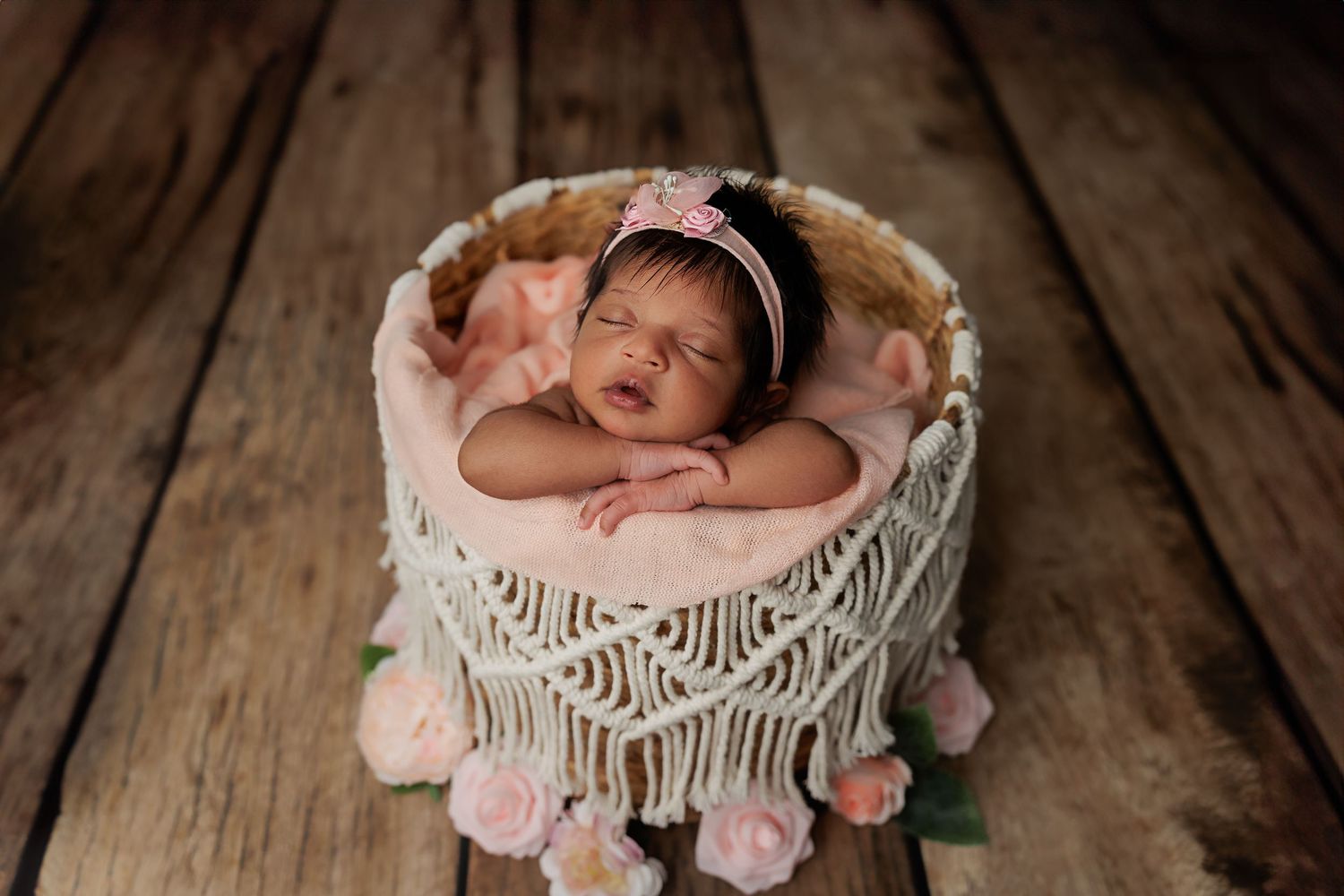 A sleeping newborn rests peacefully in a macrame basket decorated with pink pom poms on a wooden floor.