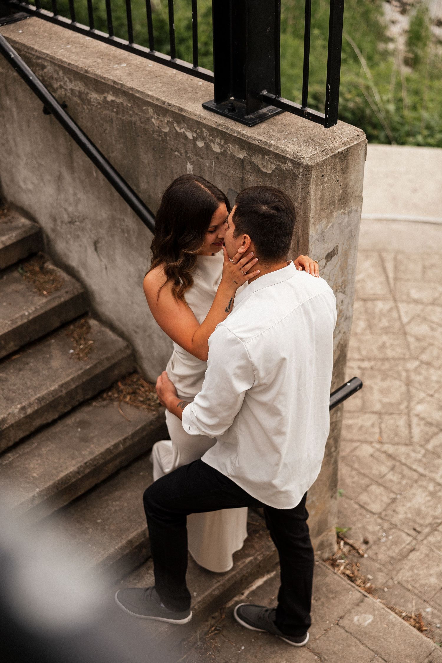 Man proposing on one knee to woman on outdoor steps, both dressed in white, sharing a romantic moment.