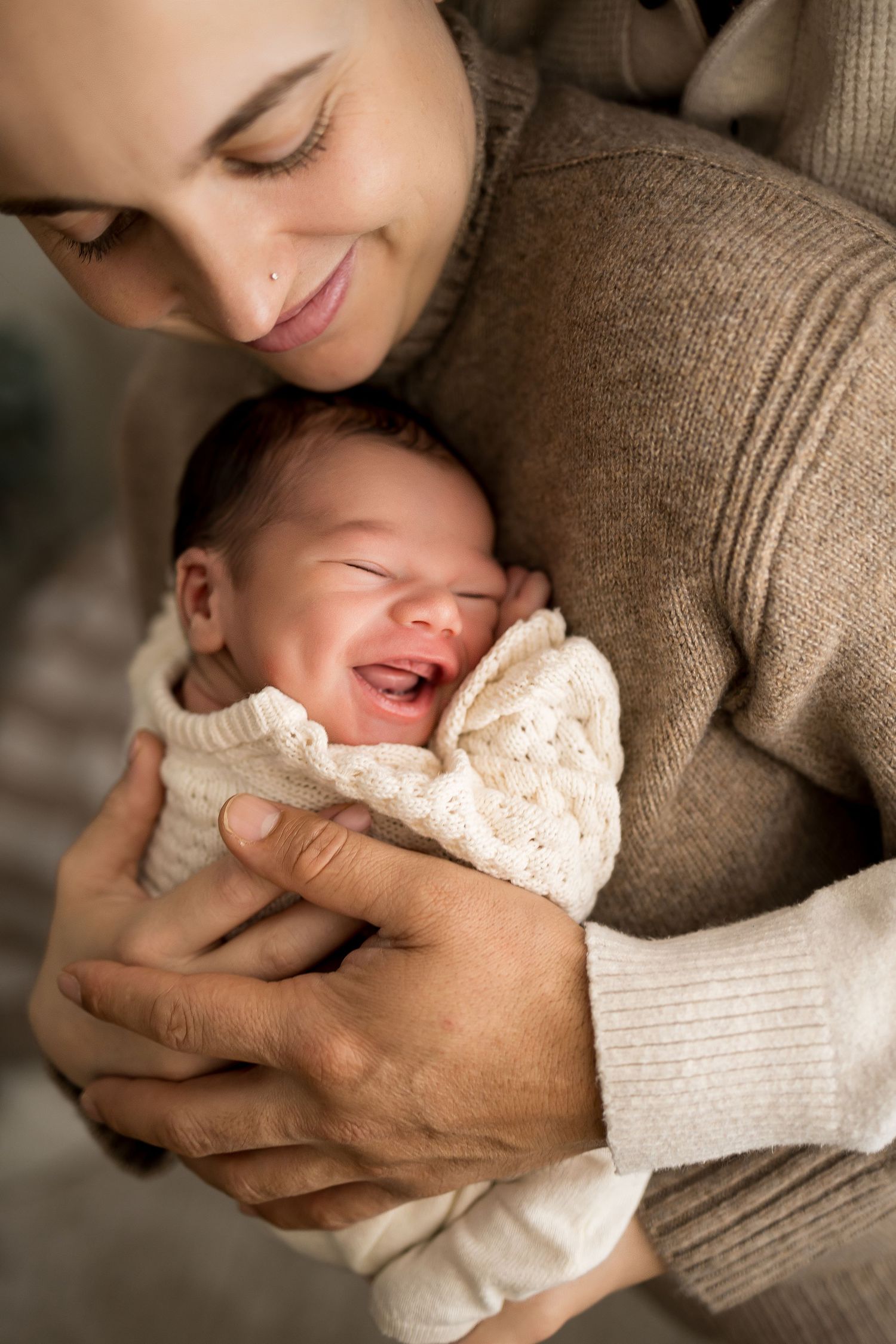 A smiling parent cradles a crying newborn baby wrapped in a white knit blanket, sharing a tender moment.