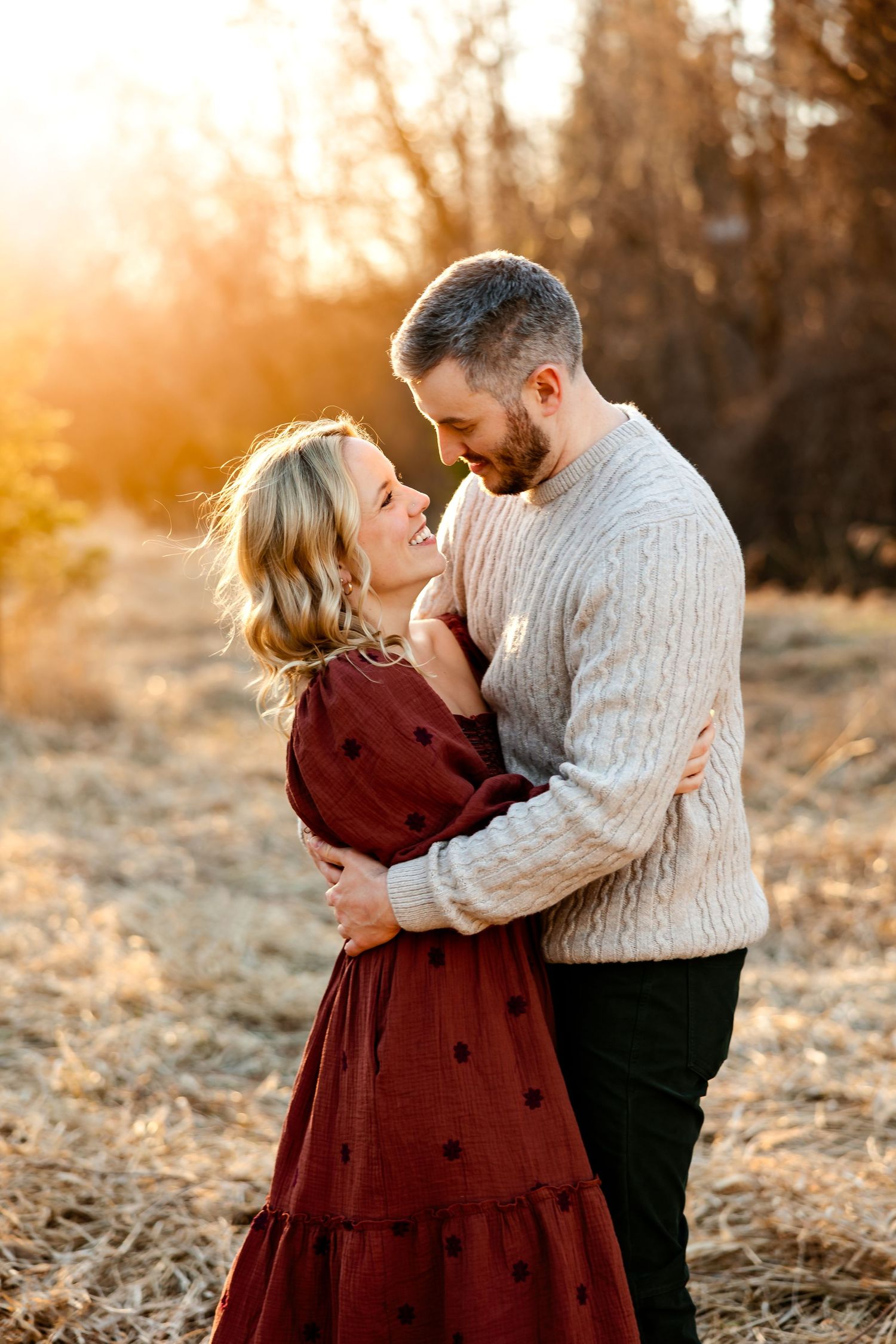 A romantic couple embraces in a field during golden hour wearing burgundy and cream colored sweaters.