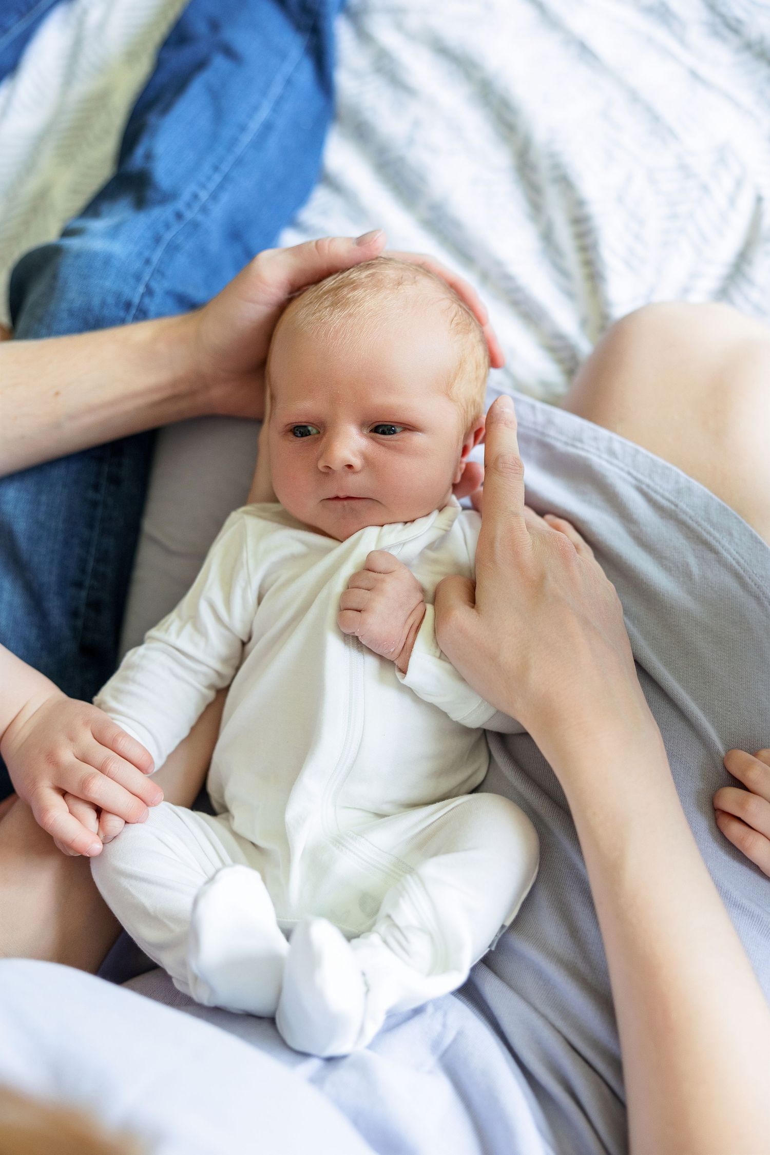 A small baby wearing a white onesie lays on soft bedding while gentle hands provide comforting touches.