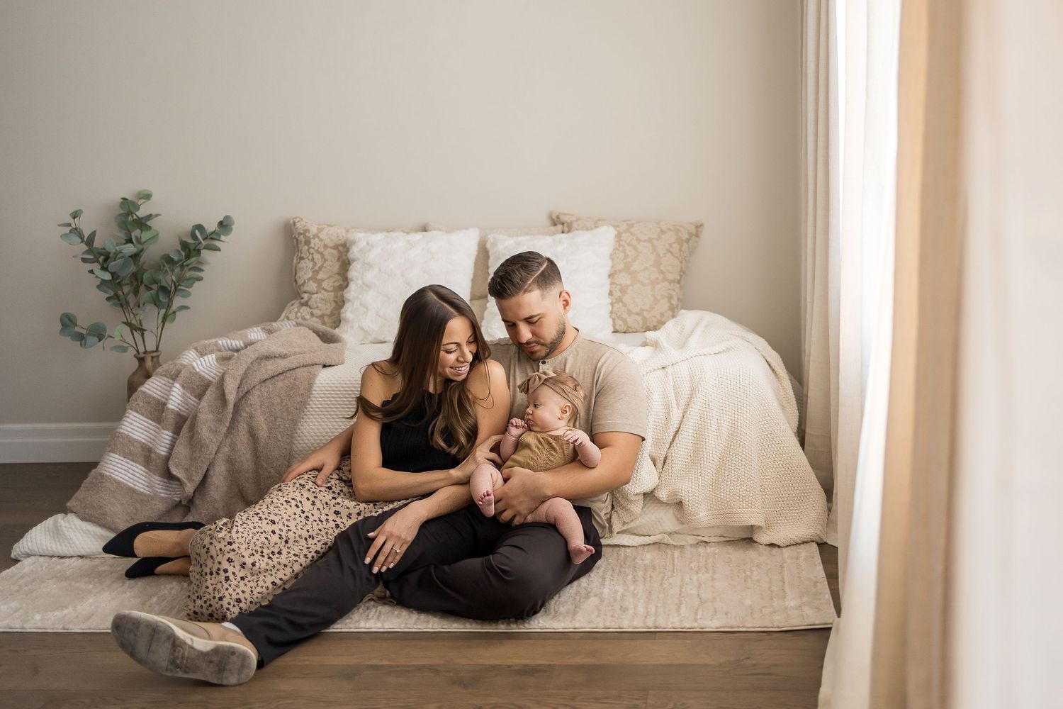 A young family sits on the floor by a bed, parents smiling at their newborn baby in a bright, cozy bedroom.