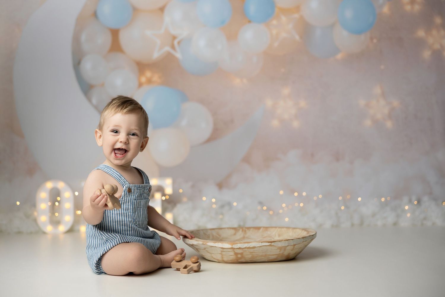 A baby lying on a gray backdrop near pastel balloons and a wooden bowl in a dreamy first birthday photo setup.