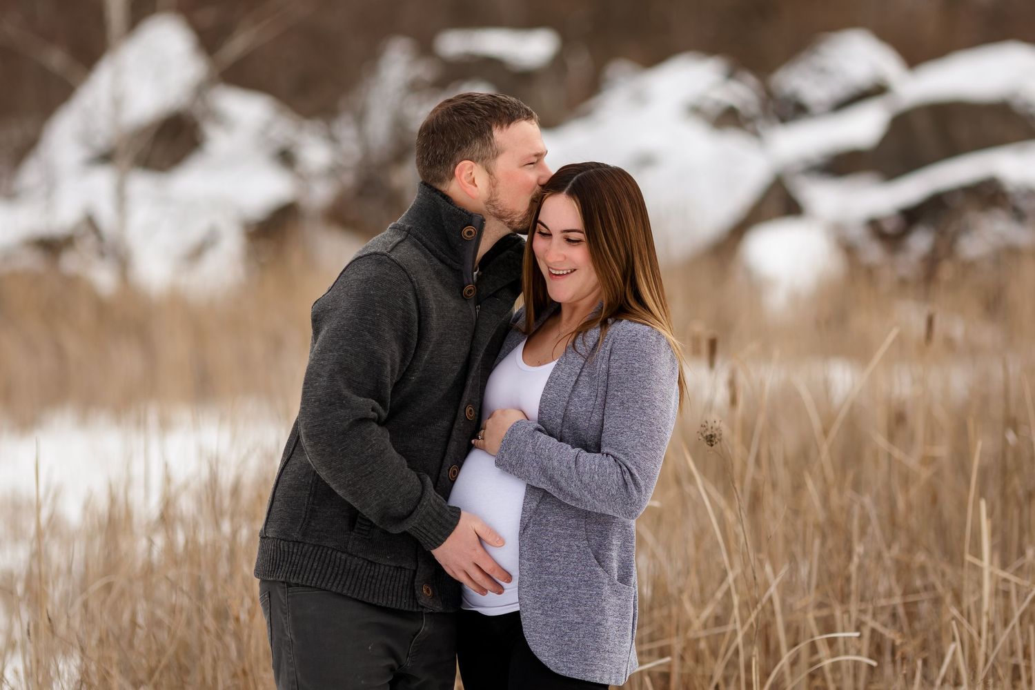 Winter maternity photo of couple in warm sweaters standing among snowy scenery.