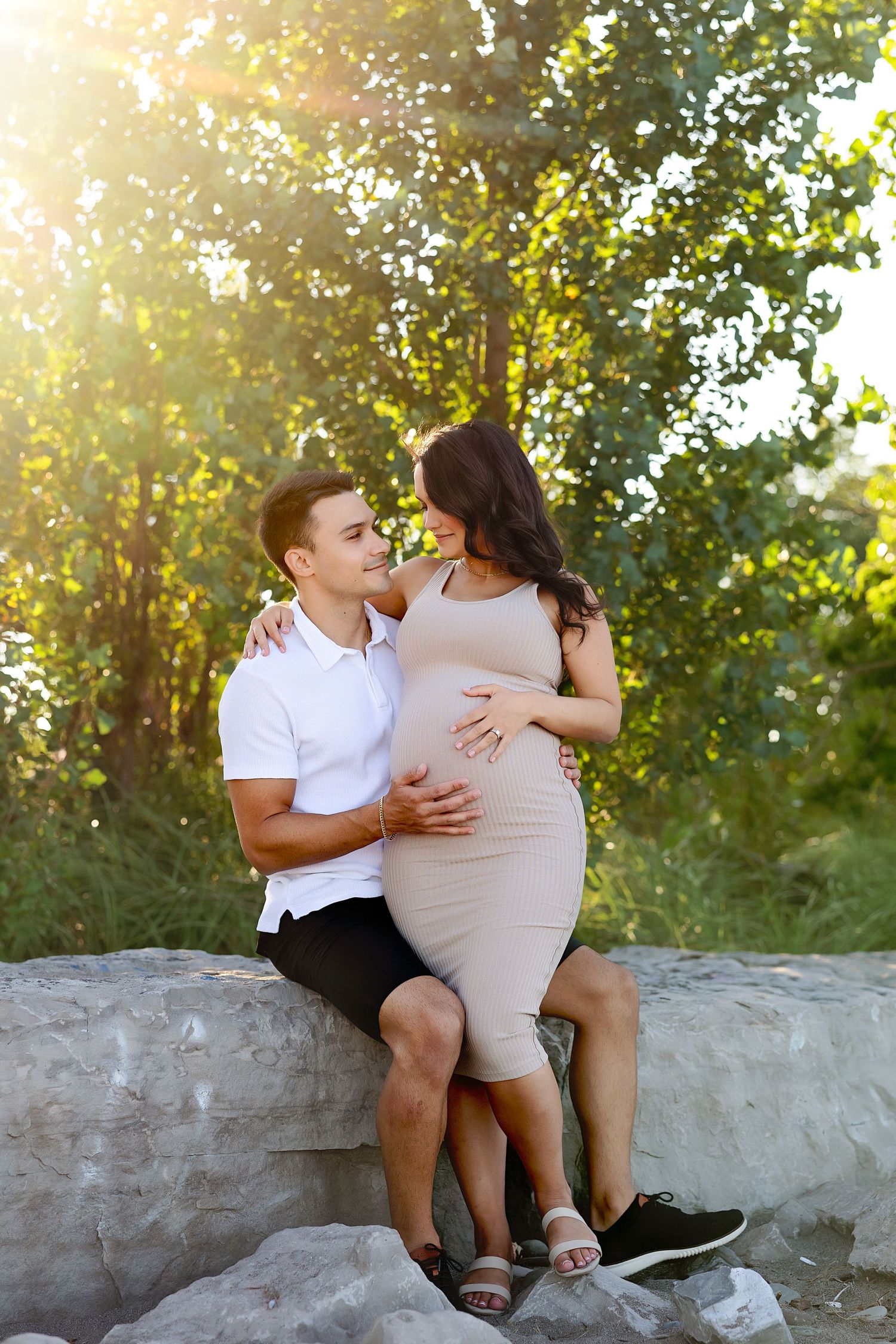 Expecting couple shares intimate moment while sitting on rock during golden hour photoshoot.