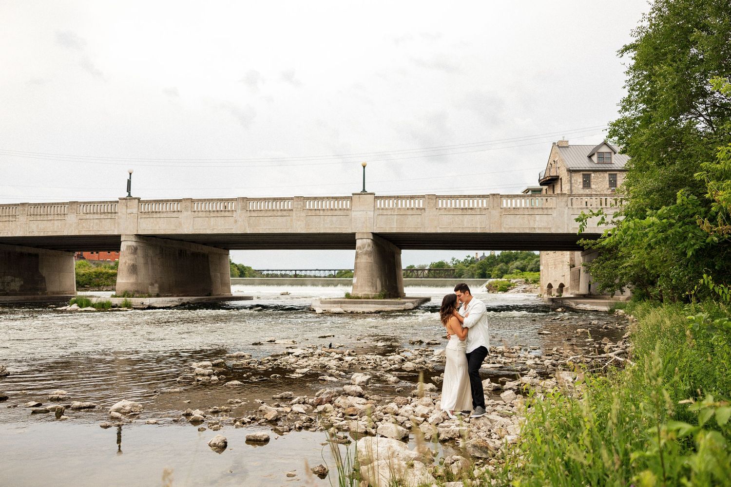 Bride and groom embrace on rocky riverbank beneath a stone bridge on a cloudy day.