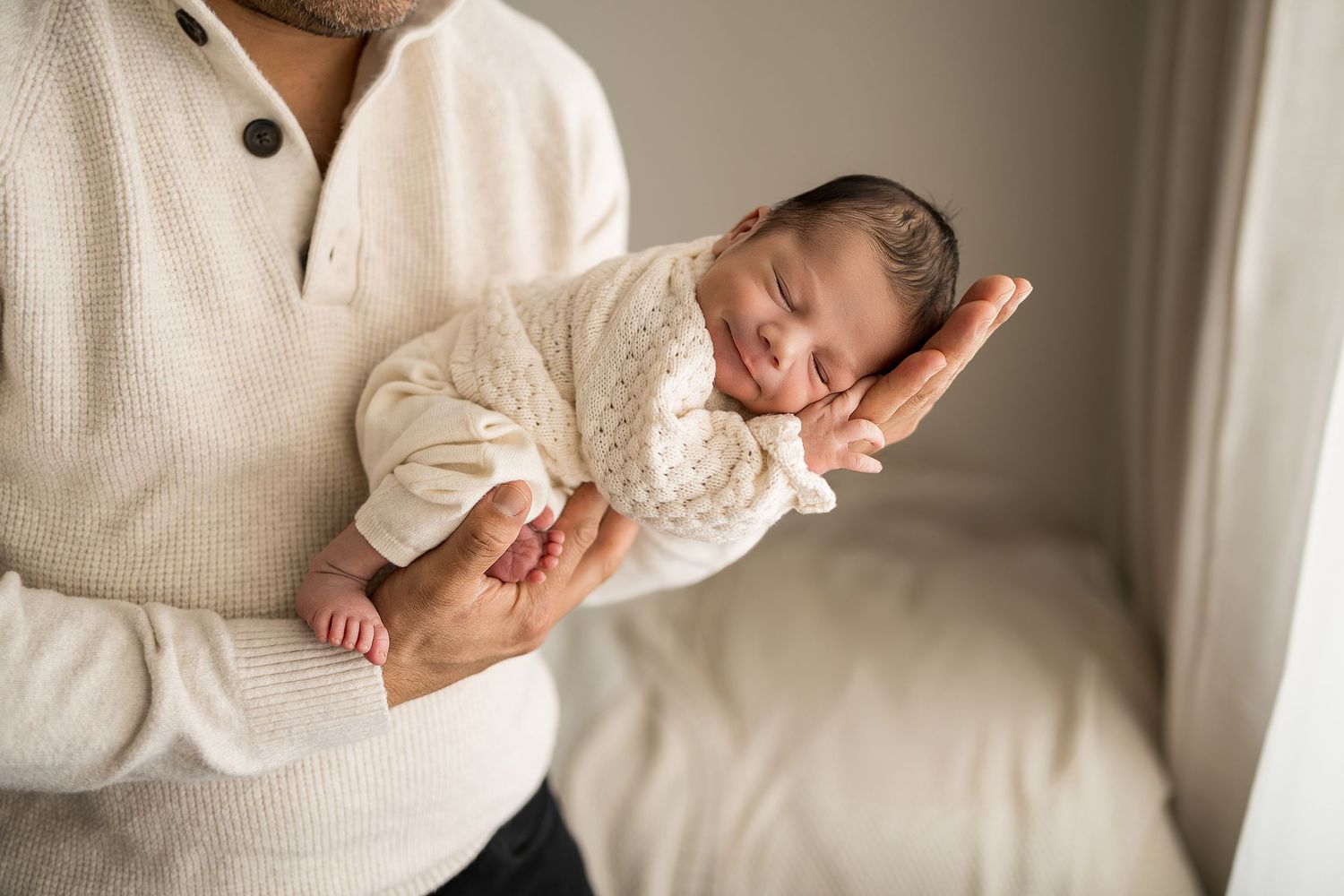 A father in a cream sweater gently holds his sleeping newborn baby dressed in a white knit outfit.