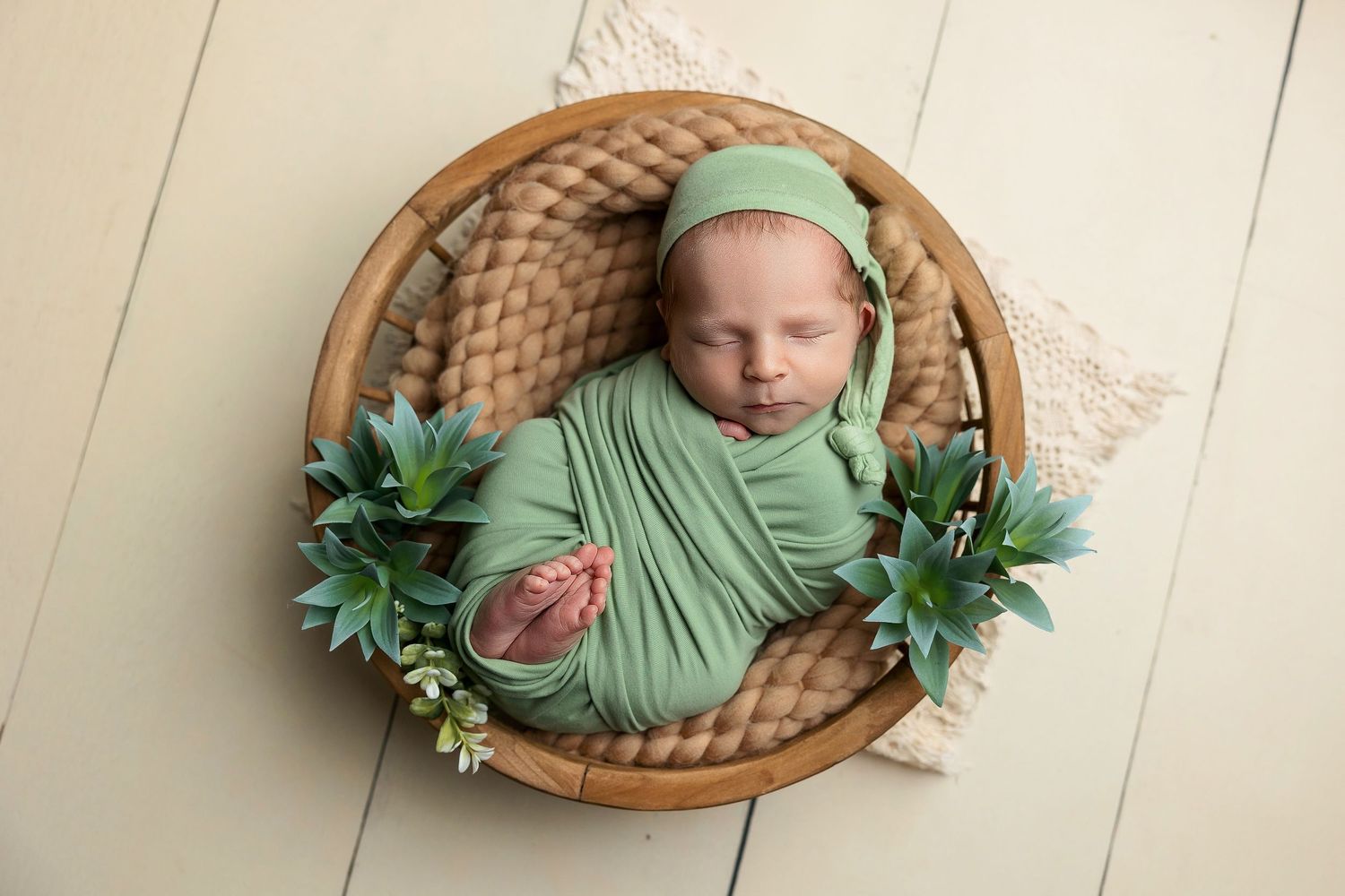 Sleeping newborn wrapped in soft green fabric lying in woven basket with decorative greenery.