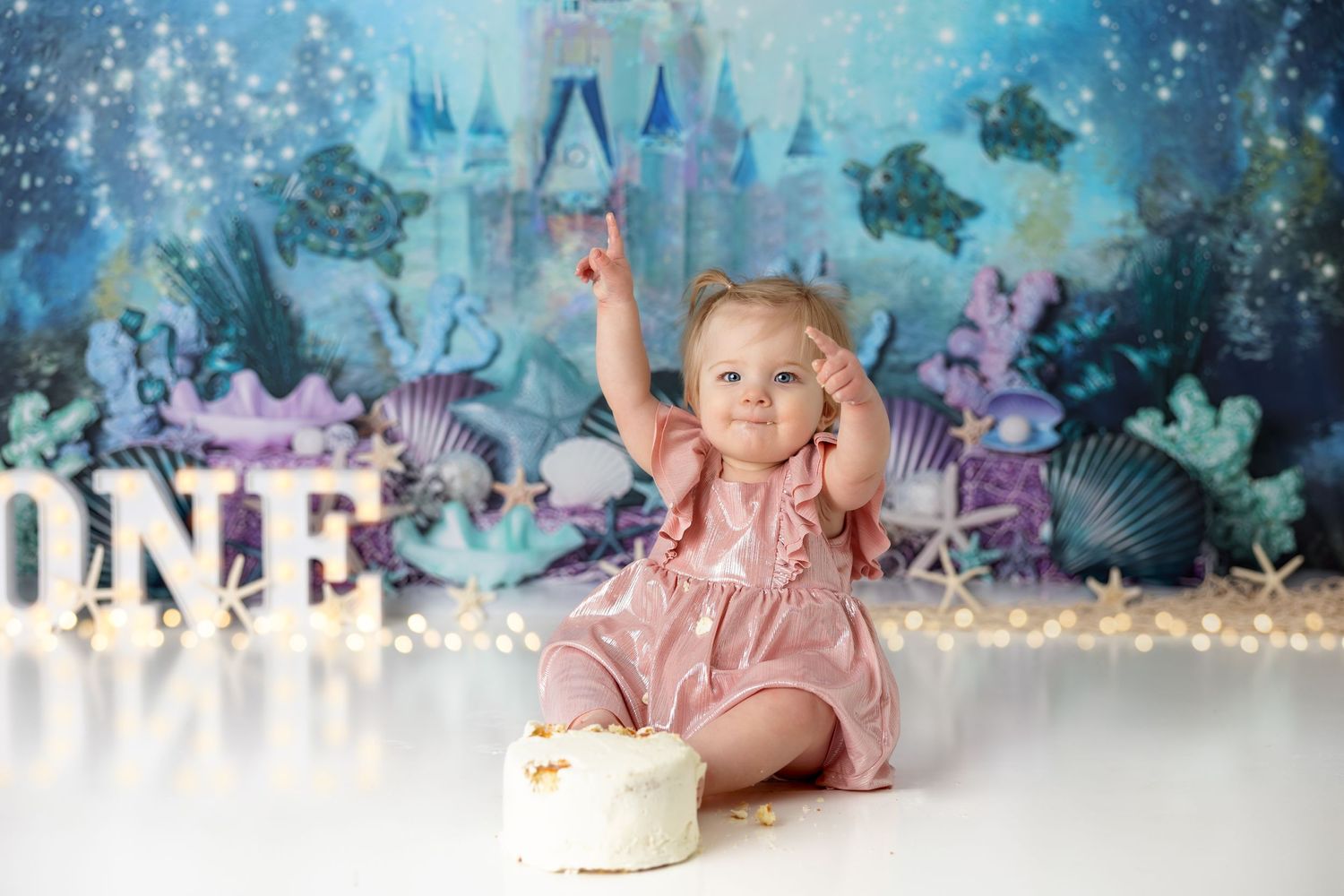 Excited baby in pink dress reaches up during underwater themed first birthday photo shoot.