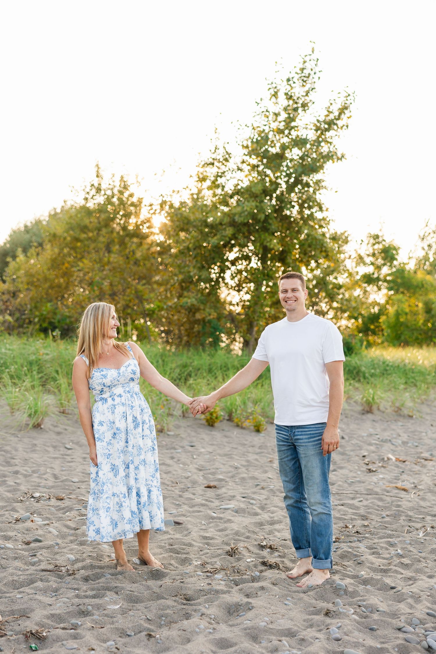 Couple holding hands while standing barefoot on sandy beach at sunset with trees in background.