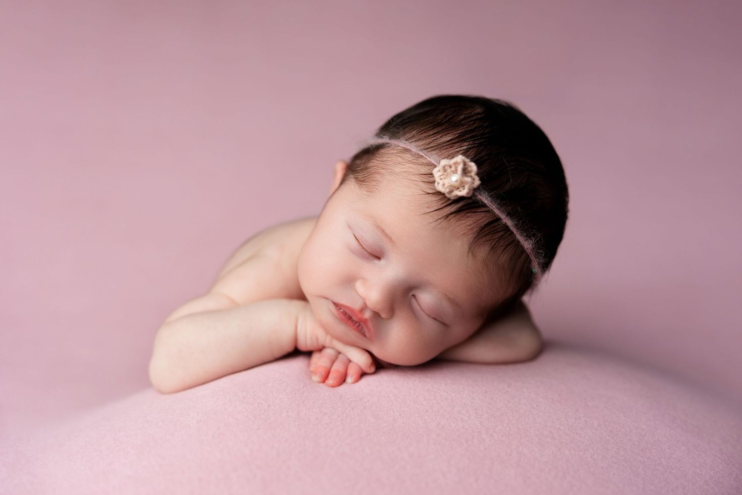 A sleeping newborn with a delicate headpiece rests on a pink surface.