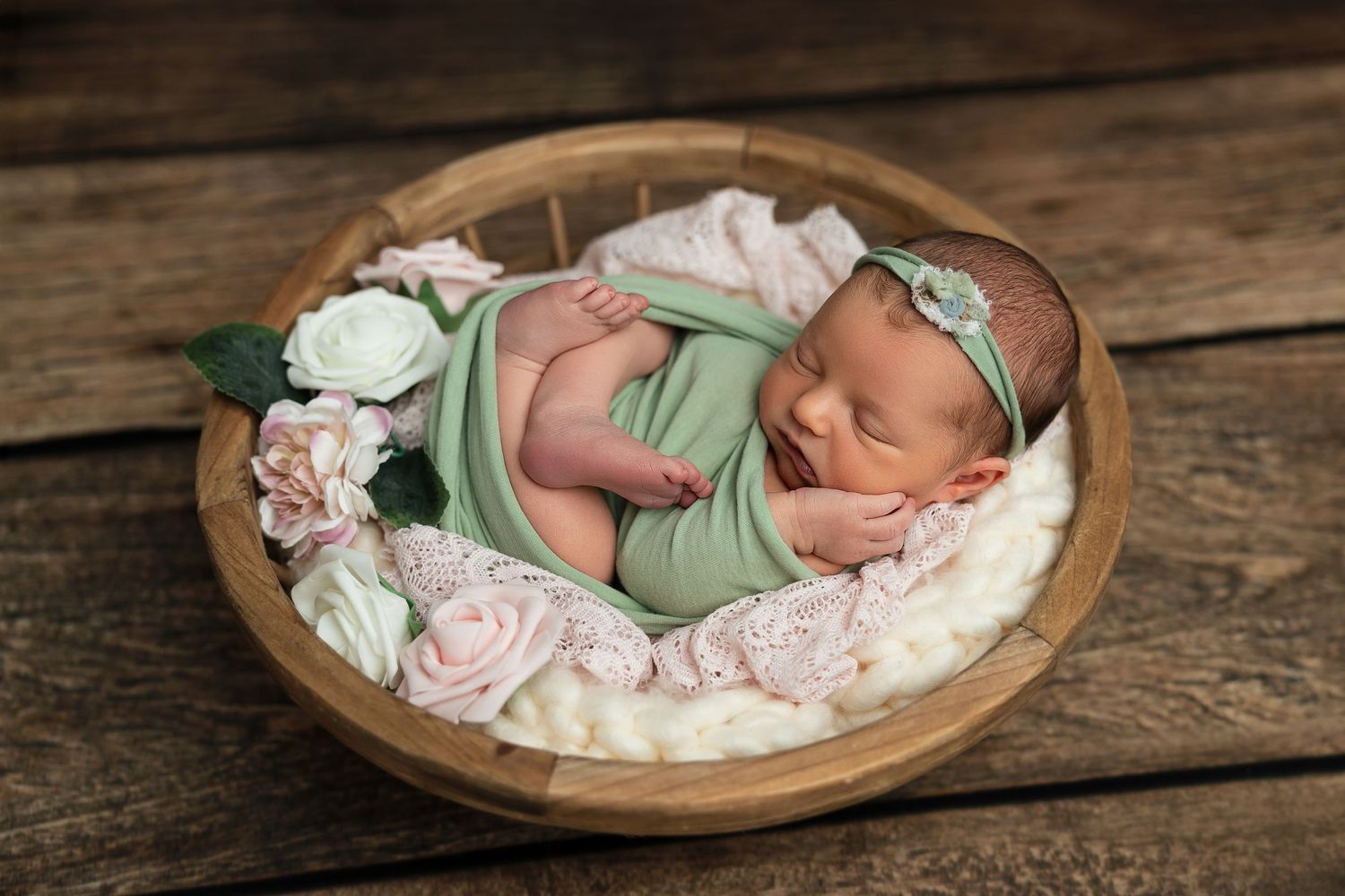 A sleeping newborn baby wearing mint green swaddle rests peacefully in a rustic wooden bowl on weathered boards.